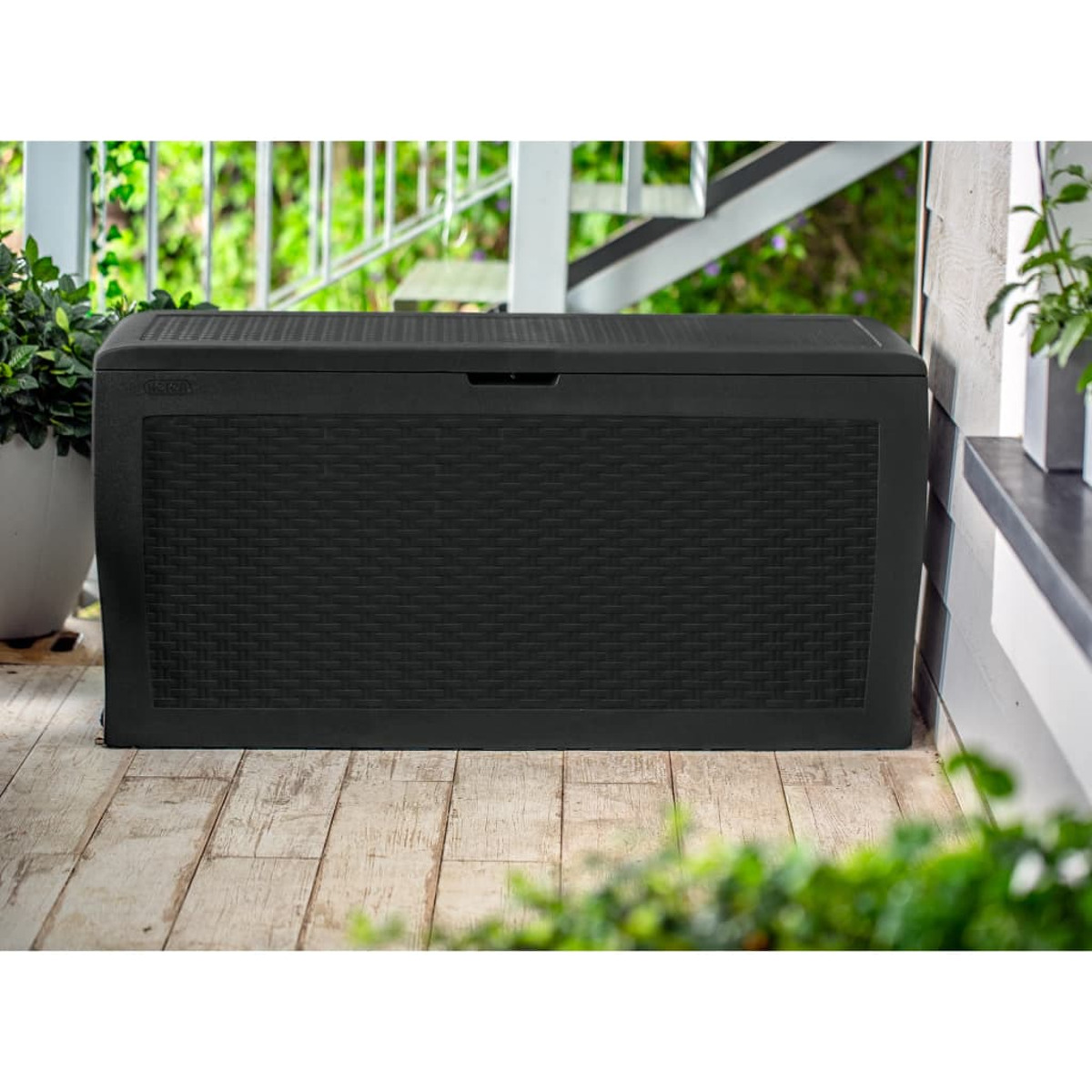 Schwarze Outdoor-Aufbewahrungsbox aus Korbgeflecht, auf Holzterrasse mit Pflanzen.