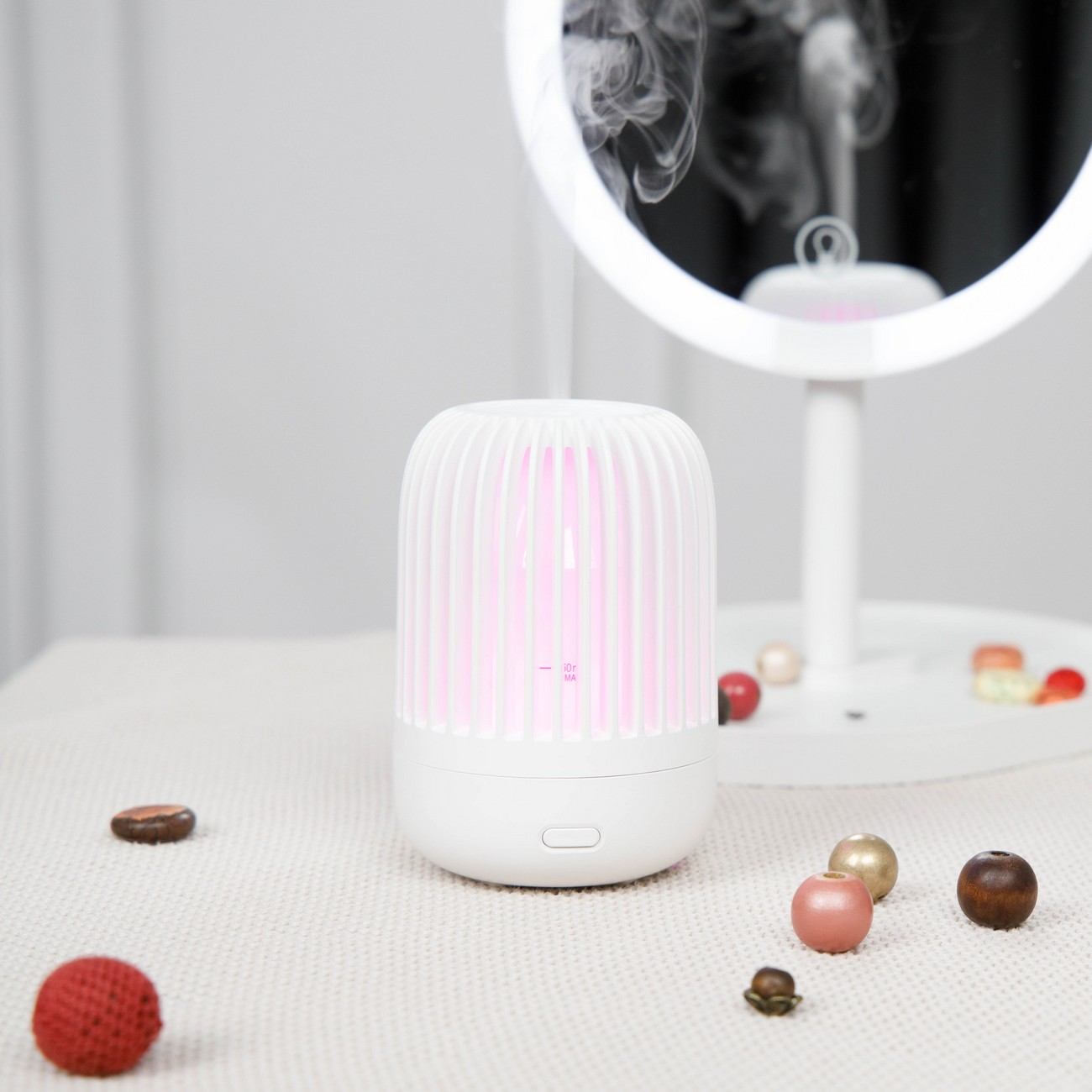Un difusor de aceite esencial blanco con luz rosa, rodeado de cuentas y un espejo. Una mesa de madera es la superficie.