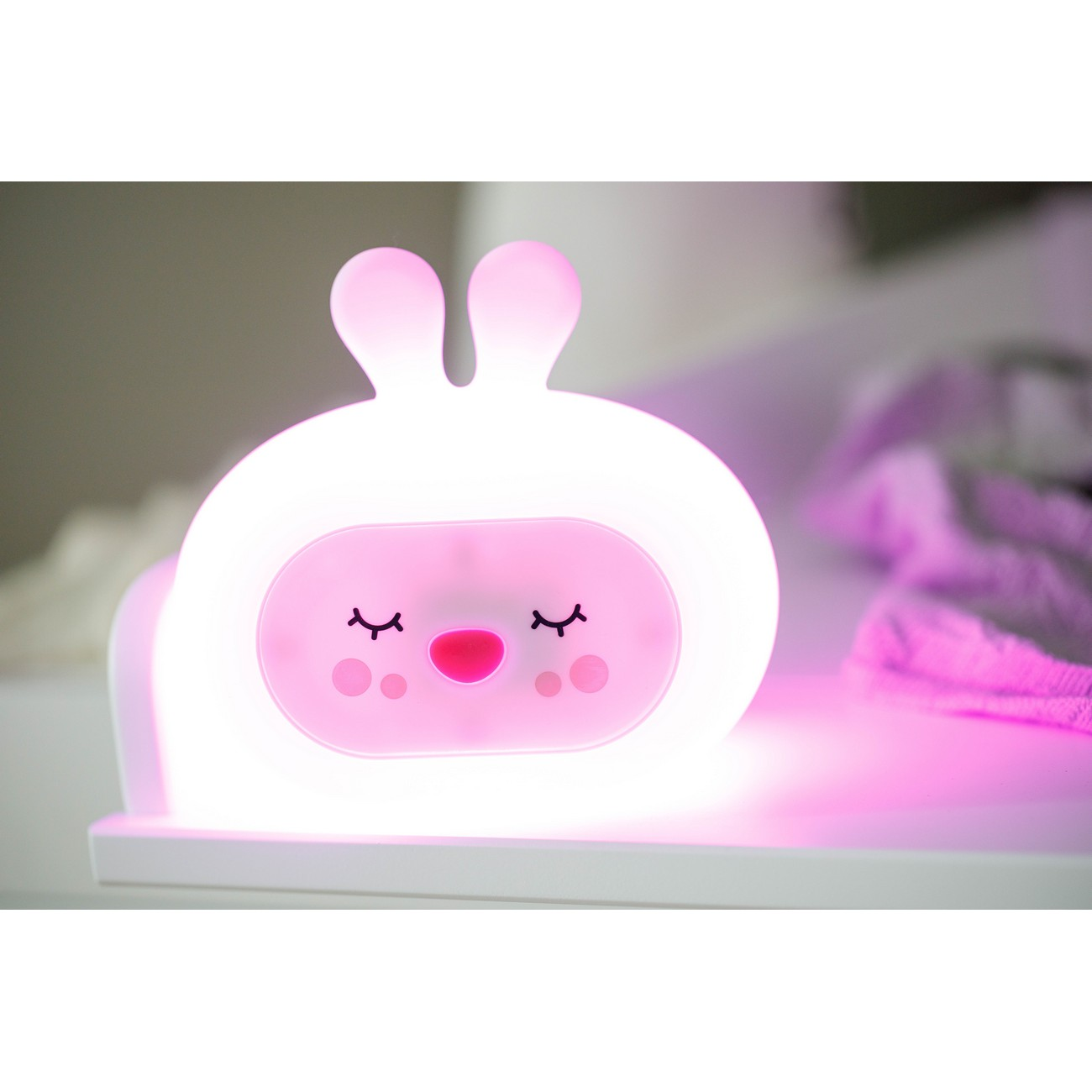 Una luz de noche con forma de conejito blanco y rosa está sobre una estantería blanca con brillo rosa.