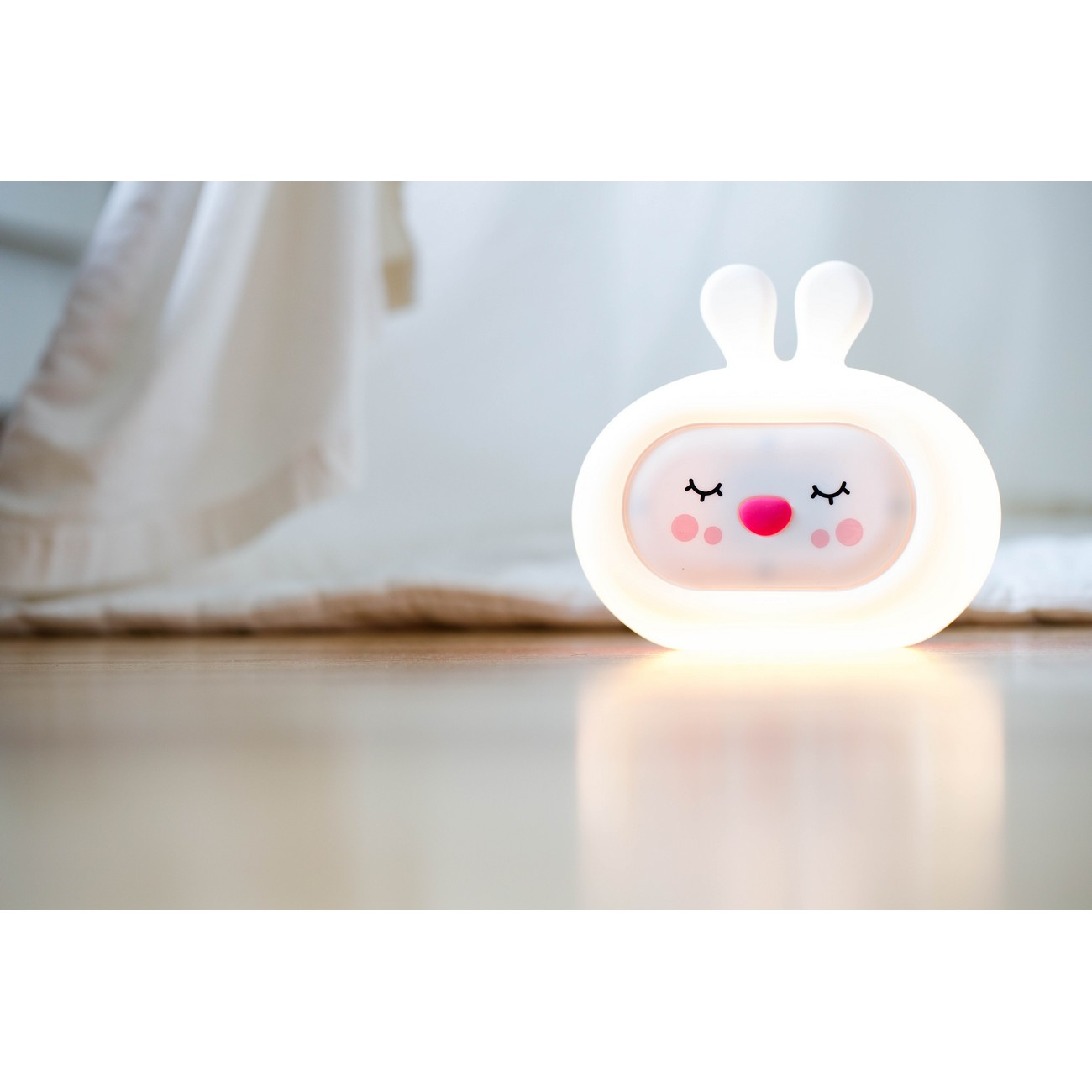 Una luz nocturna blanca en forma de conejito con ojos cerrados y nariz rosa en suelo de madera.