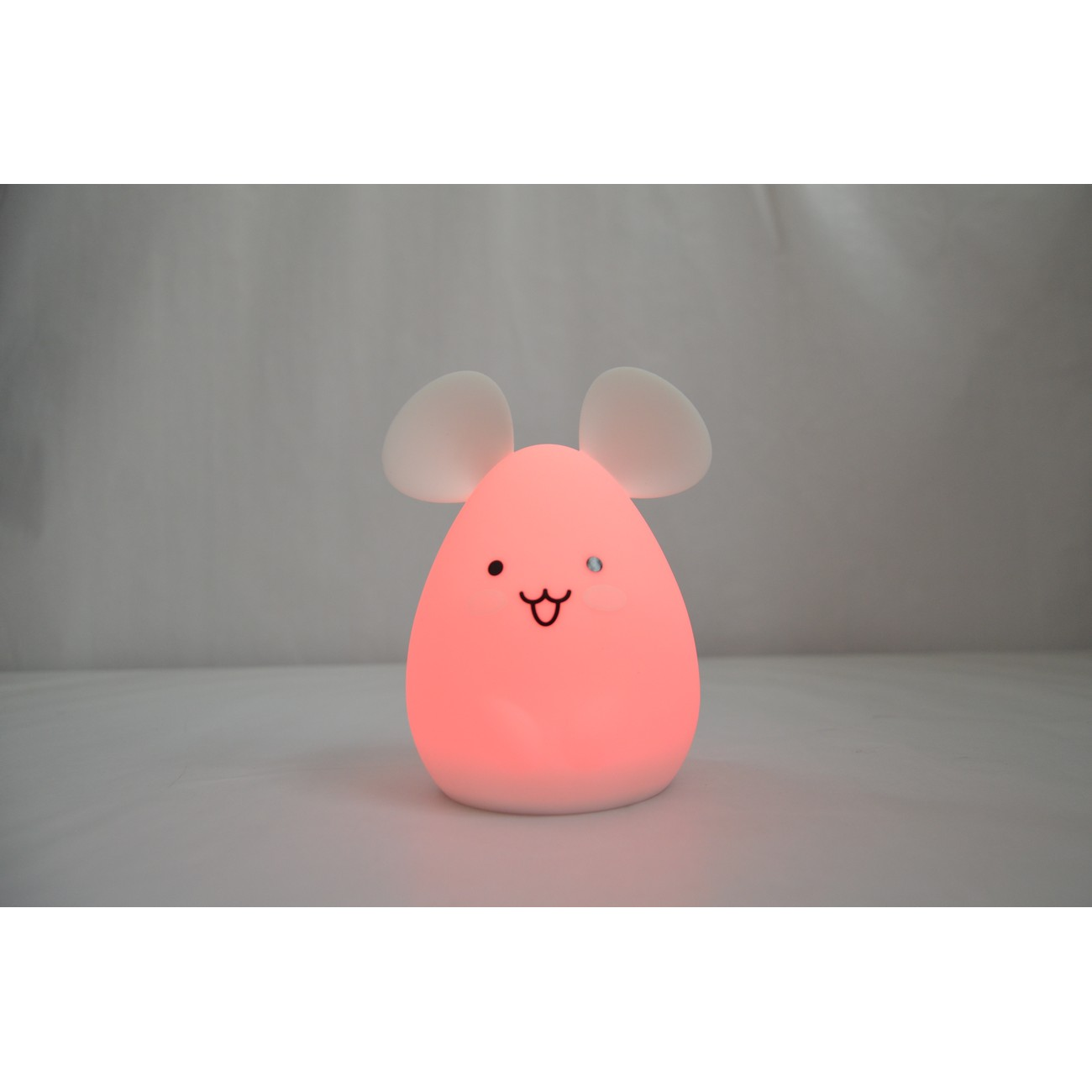 Luz de noche rosa con forma de ratón con cara sonriente y orejas blancas. Sobre una superficie blanca.