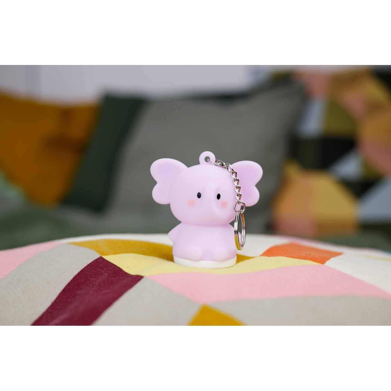 Figurita de elefante rosa sobre almohada estampada. Tiene un llavero y un anillo plateado. Fondo suave.
