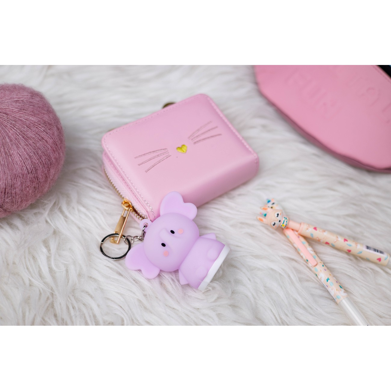 Cartera rosa con cara de gato, un elefante morado y bolígrafos sobre una superficie blanca y esponjosa.