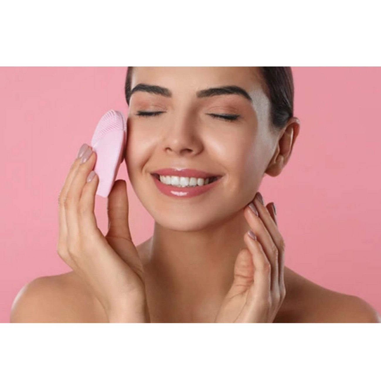 Mujer usando un cepillo de limpieza facial rosa, sonriendo, sobre un fondo rosa.