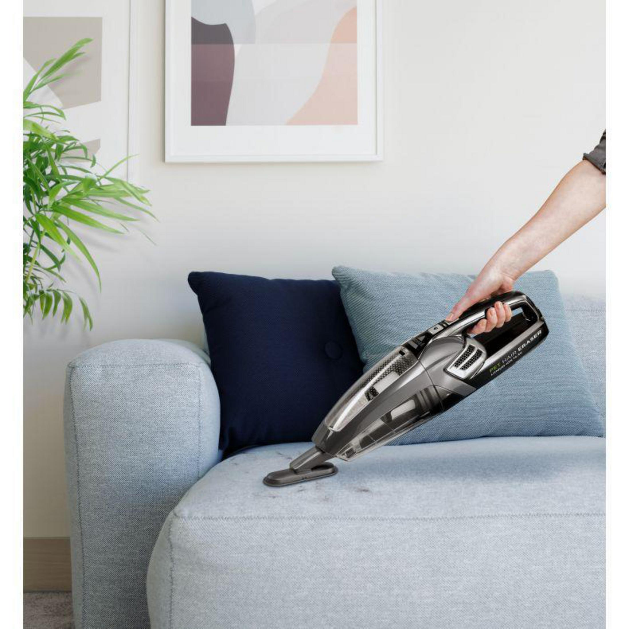 Person saugt einen hellblauen Sofa mit einem Handstaubsauger ab. Das Sofa hat ein dunkelblaues Kissen.