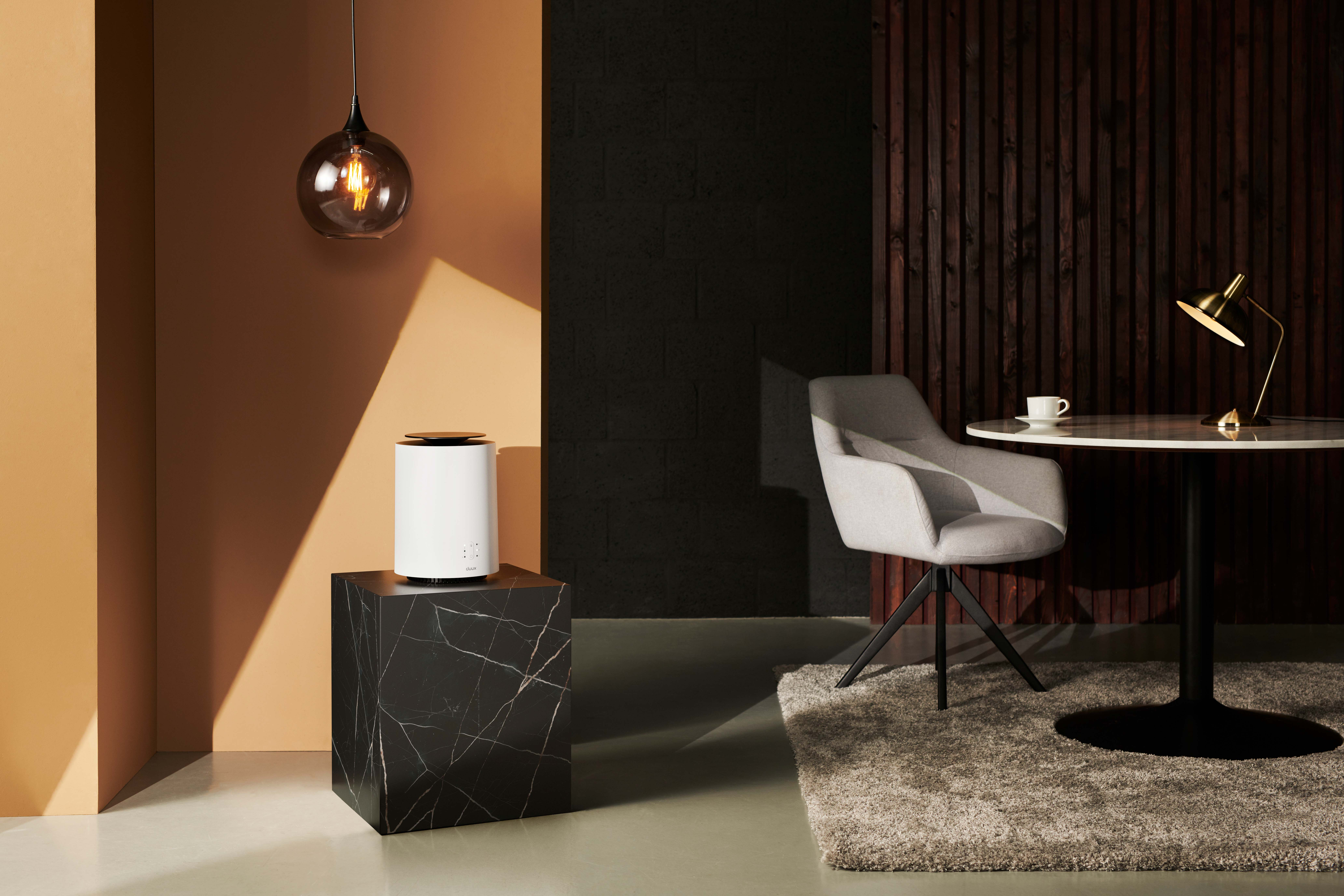 Nowoczesne wnętrze z białym nawilżaczem na czarnej kostce, szarym krzesłem i okrągłym stołem z lampą.