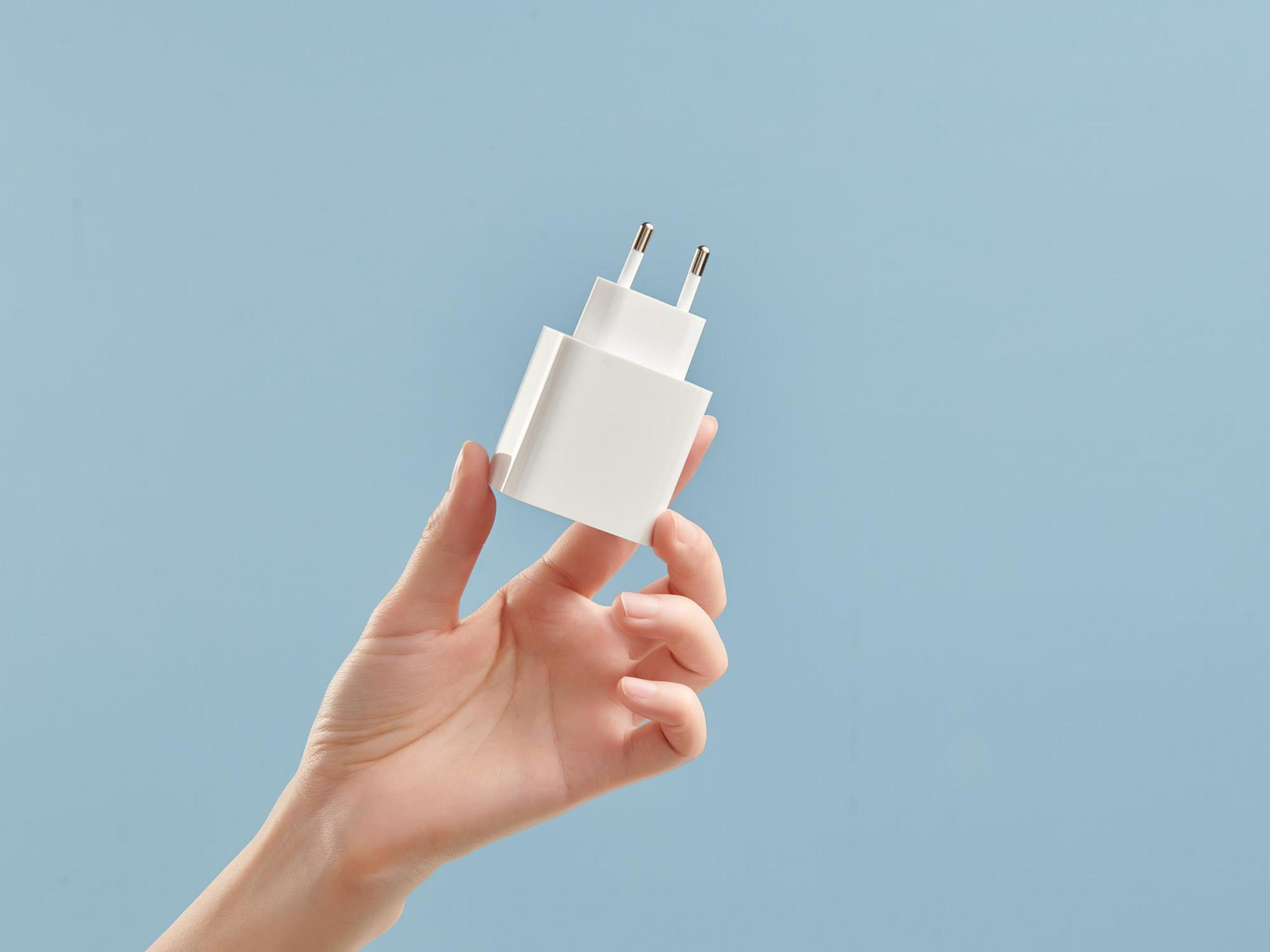 Dłoń trzyma białą ładowarkę do telefonu na jasnoniebieskim tle.