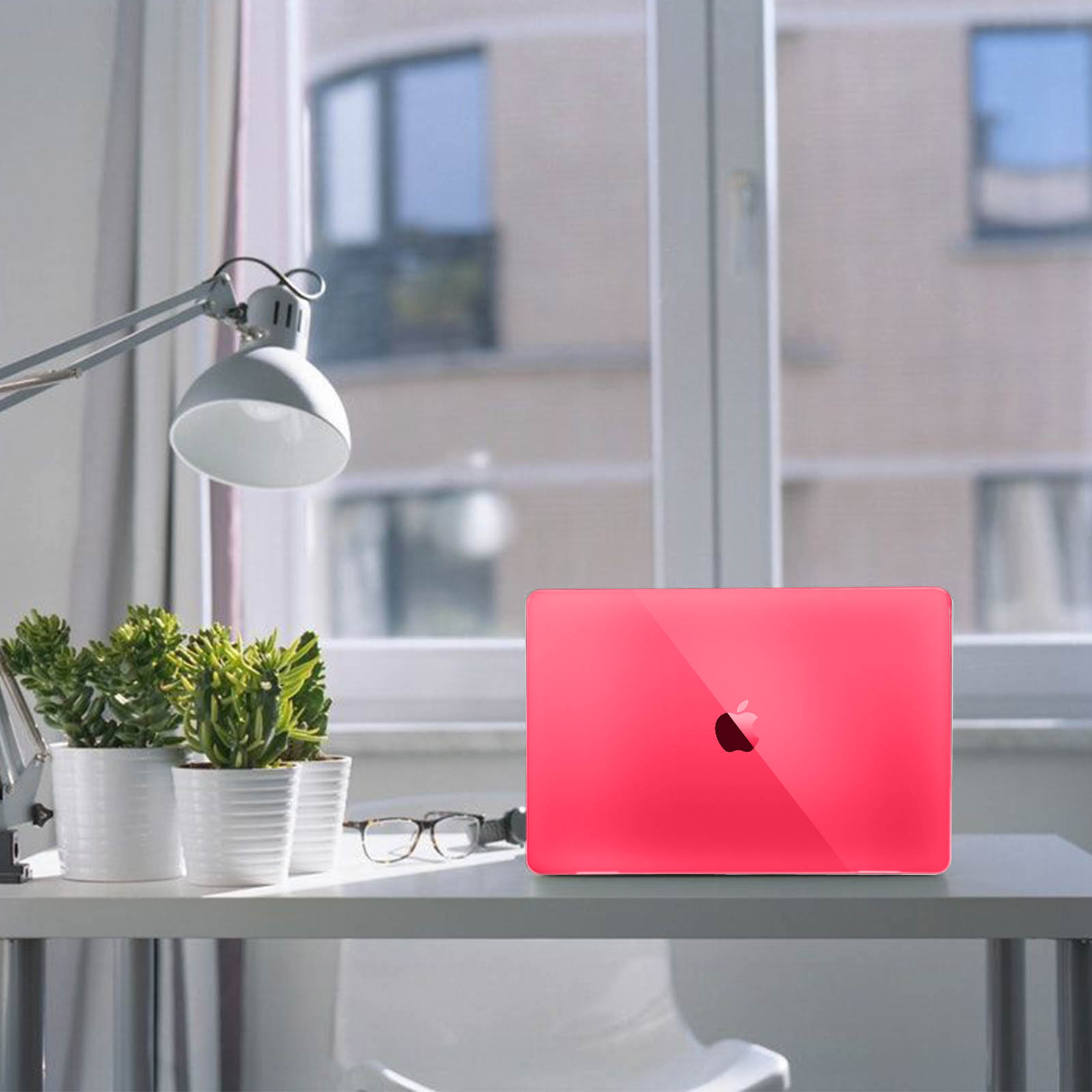 Laptop rosa sulla scrivania con piante, lampada e occhiali, vicino a una finestra.