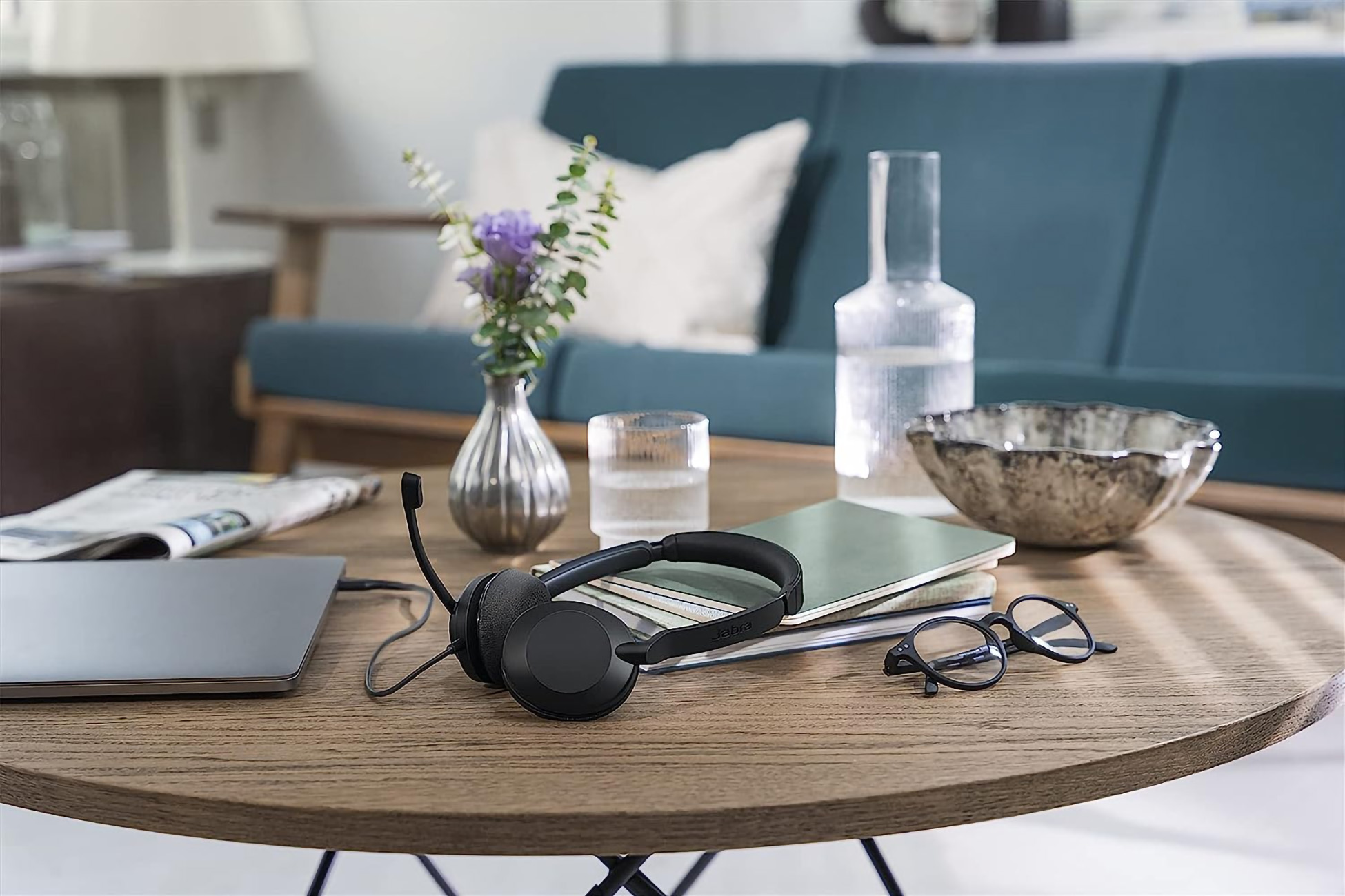 Ein Schreibtisch mit Laptop, Headset, Brille, Notizbüchern und einer Vase mit Blumen. Im Hintergrund ein Sofa.
