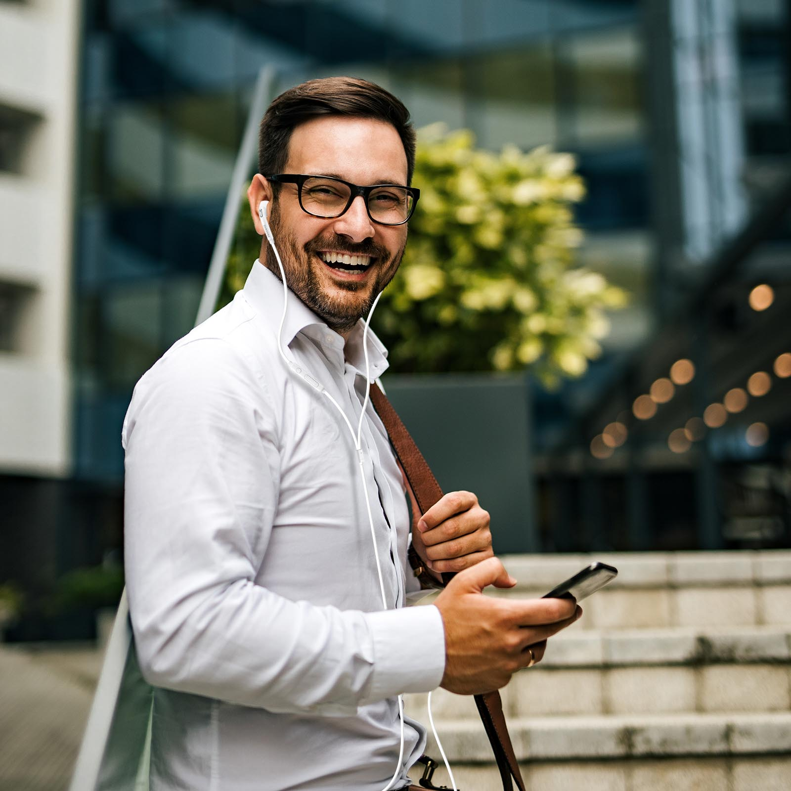 Uśmiechnięty mężczyzna w białej koszuli z okularami, trzymający telefon i torbę, ze słuchawkami, przed budynkiem.