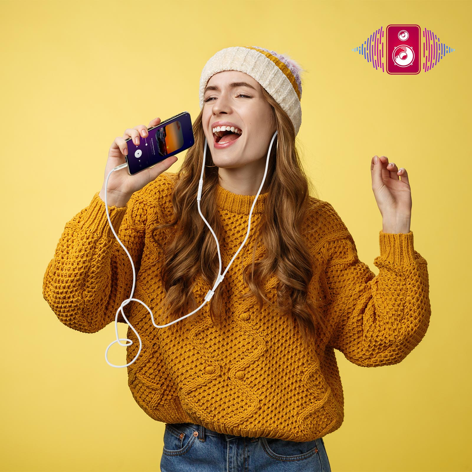 Kobieta śpiewa z telefonem. Ma na sobie żółty sweter i czapkę. Żółte tło.