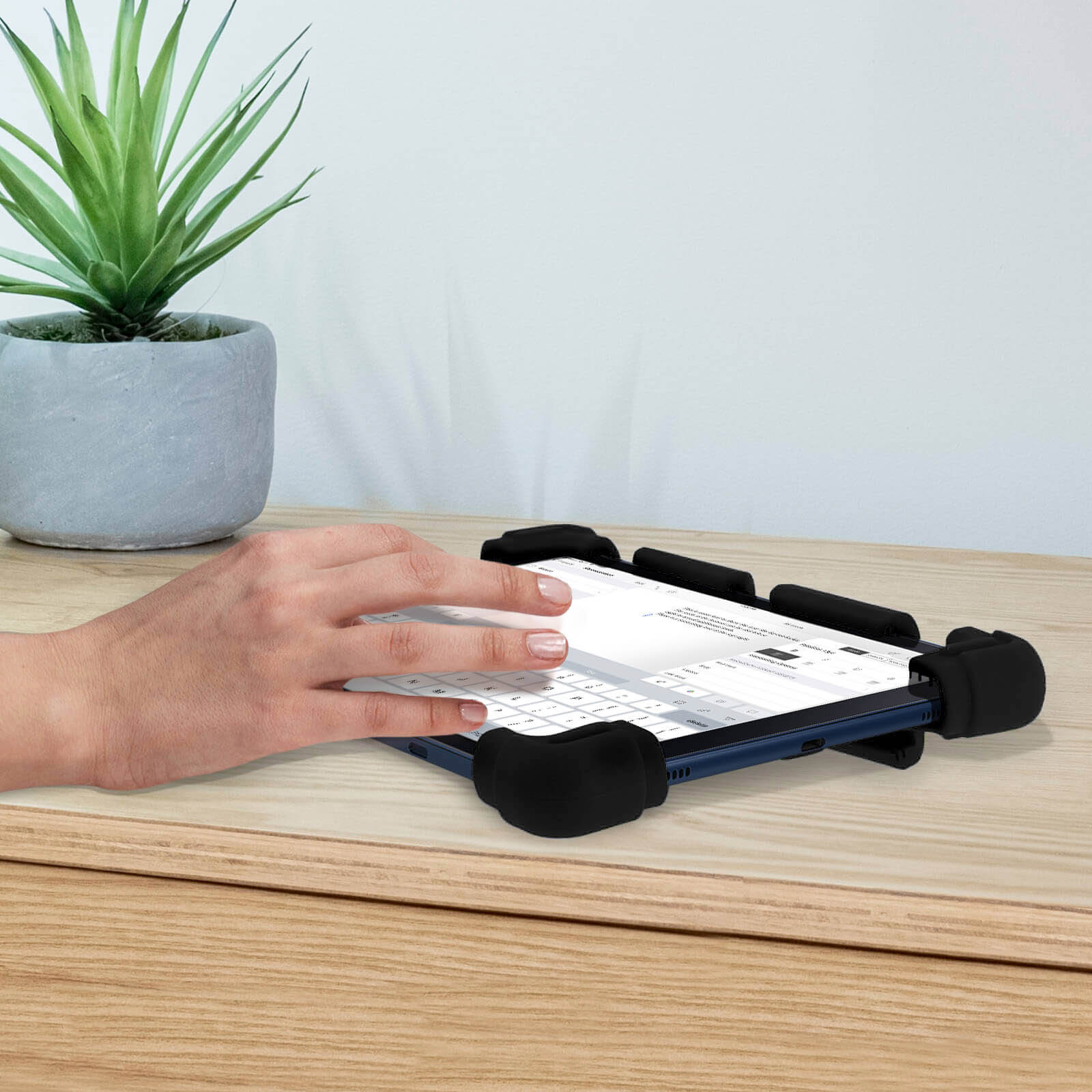 Eine Hand, die auf einem Tablet in einer schwarzen Hülle tippt, auf einem Holztisch, eine Pflanze im Hintergrund.