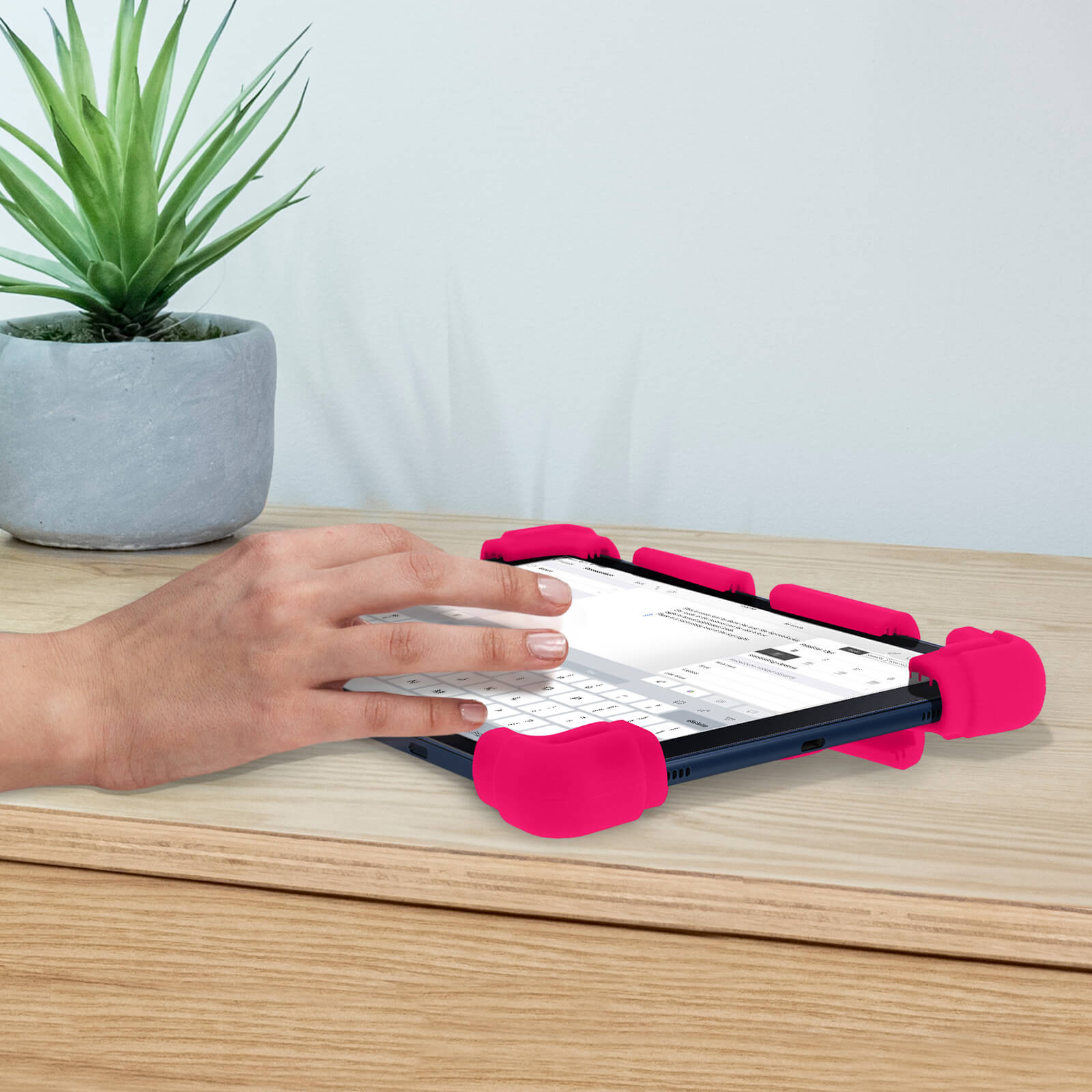 Hand auf einem Tablet in einer rosa Hülle, auf Holzoberfläche, mit Pflanze im Hintergrund.