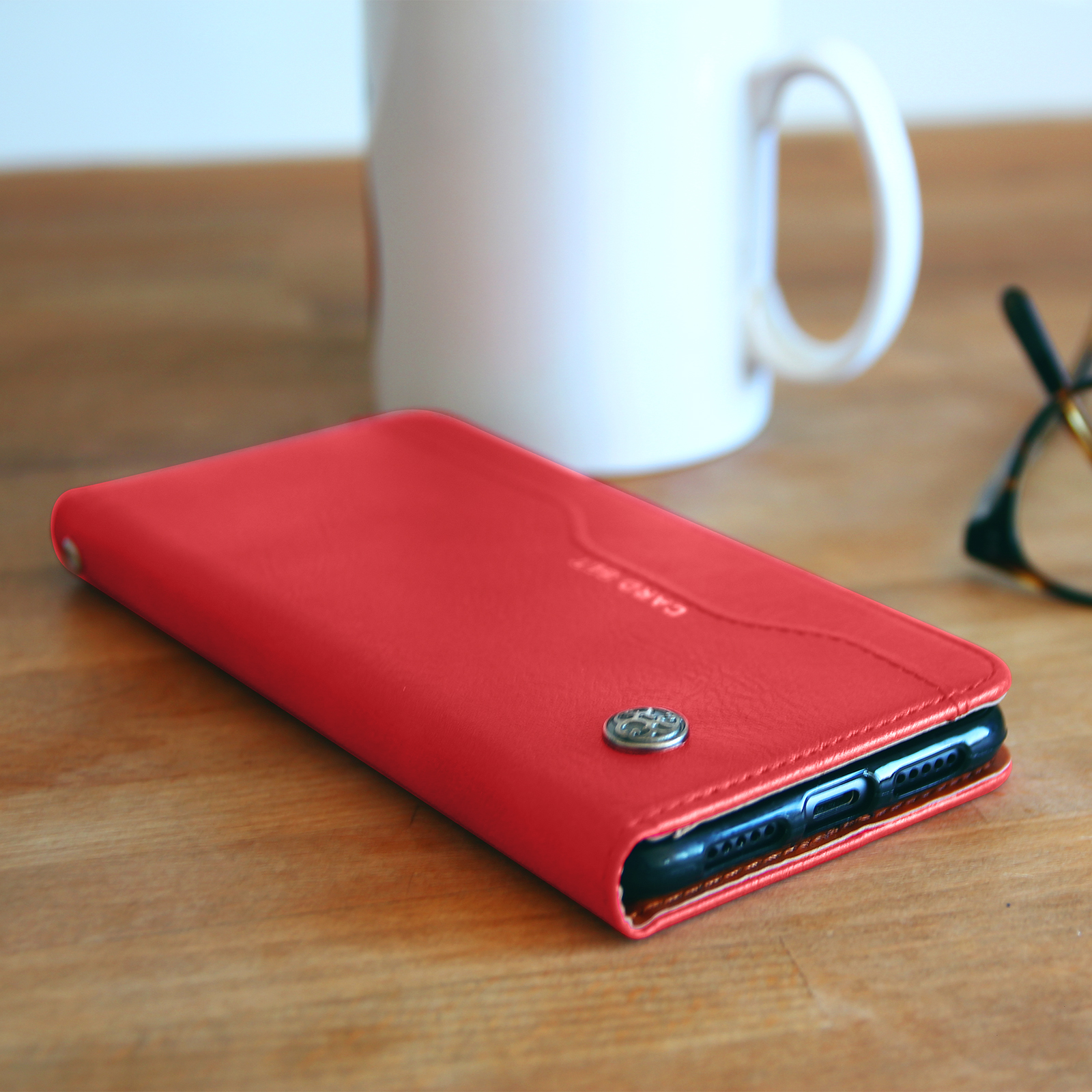 Custodia rossa con telefono, tazza bianca, occhiali su tavolo di legno.