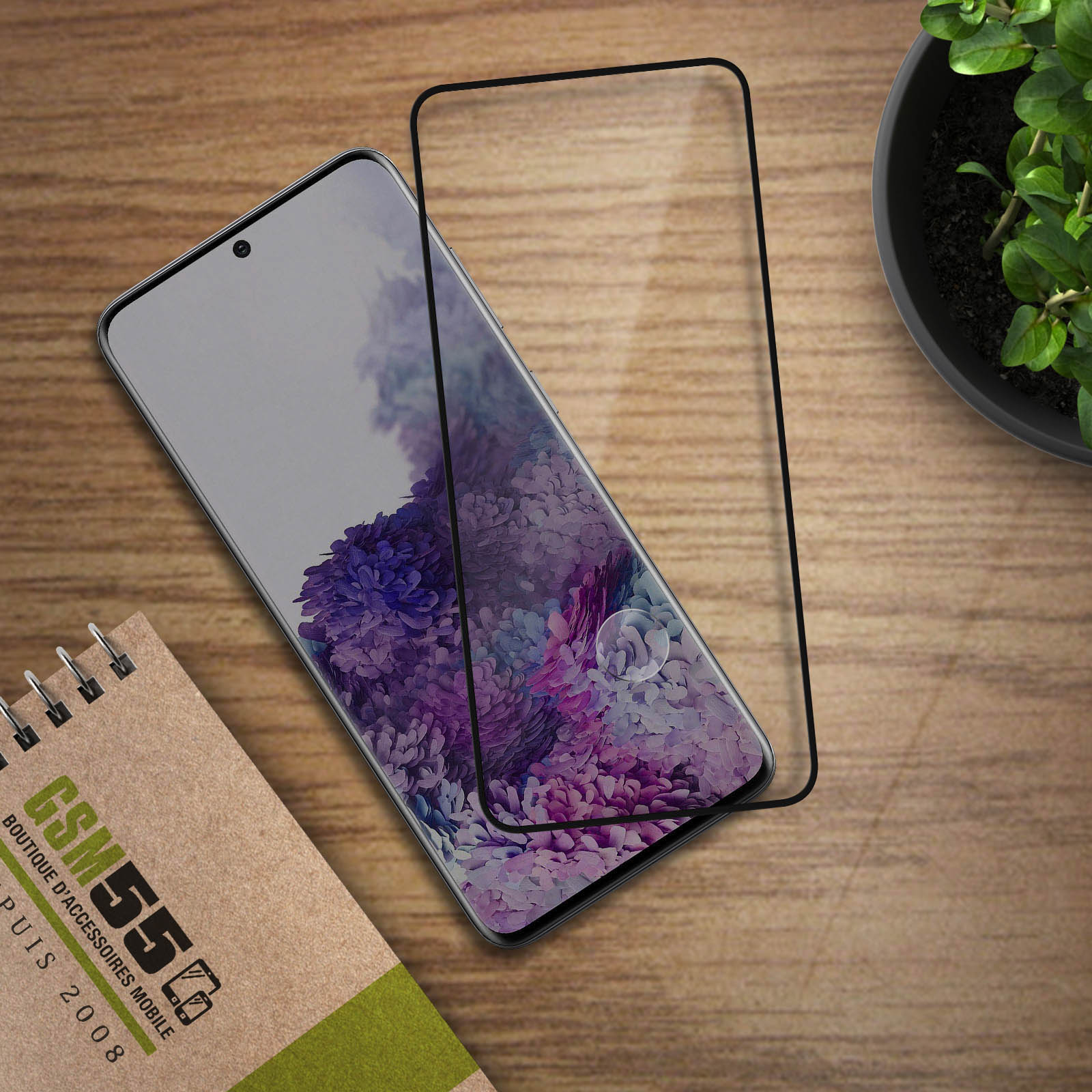 Un telefono con protezione schermo su una superficie di legno. Sono visibili anche un quaderno e una pianta.