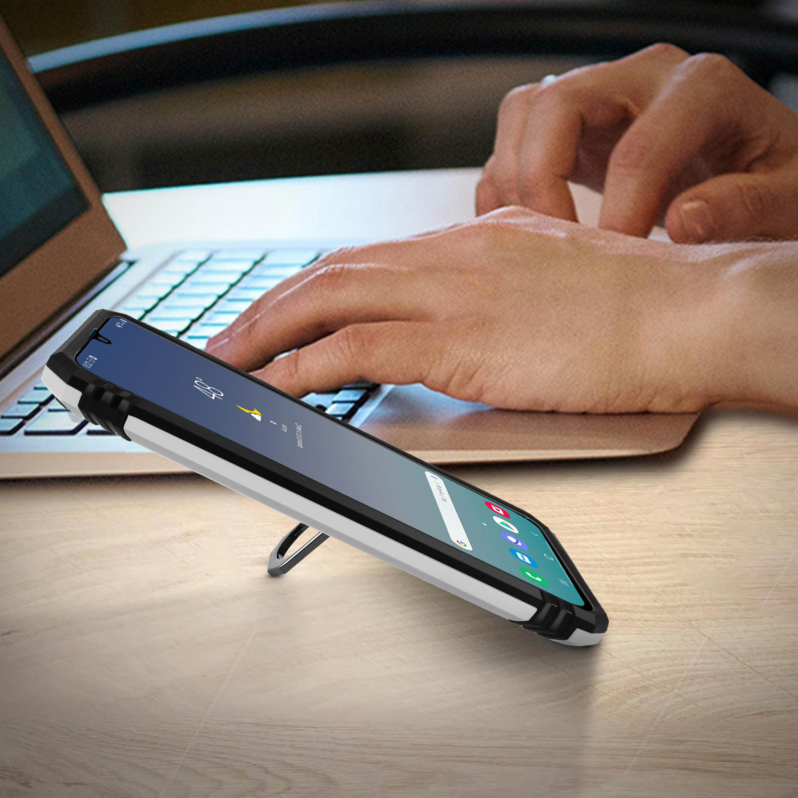 Un telefono in una custodia su un tavolo, con le mani di una persona e un laptop sullo sfondo.