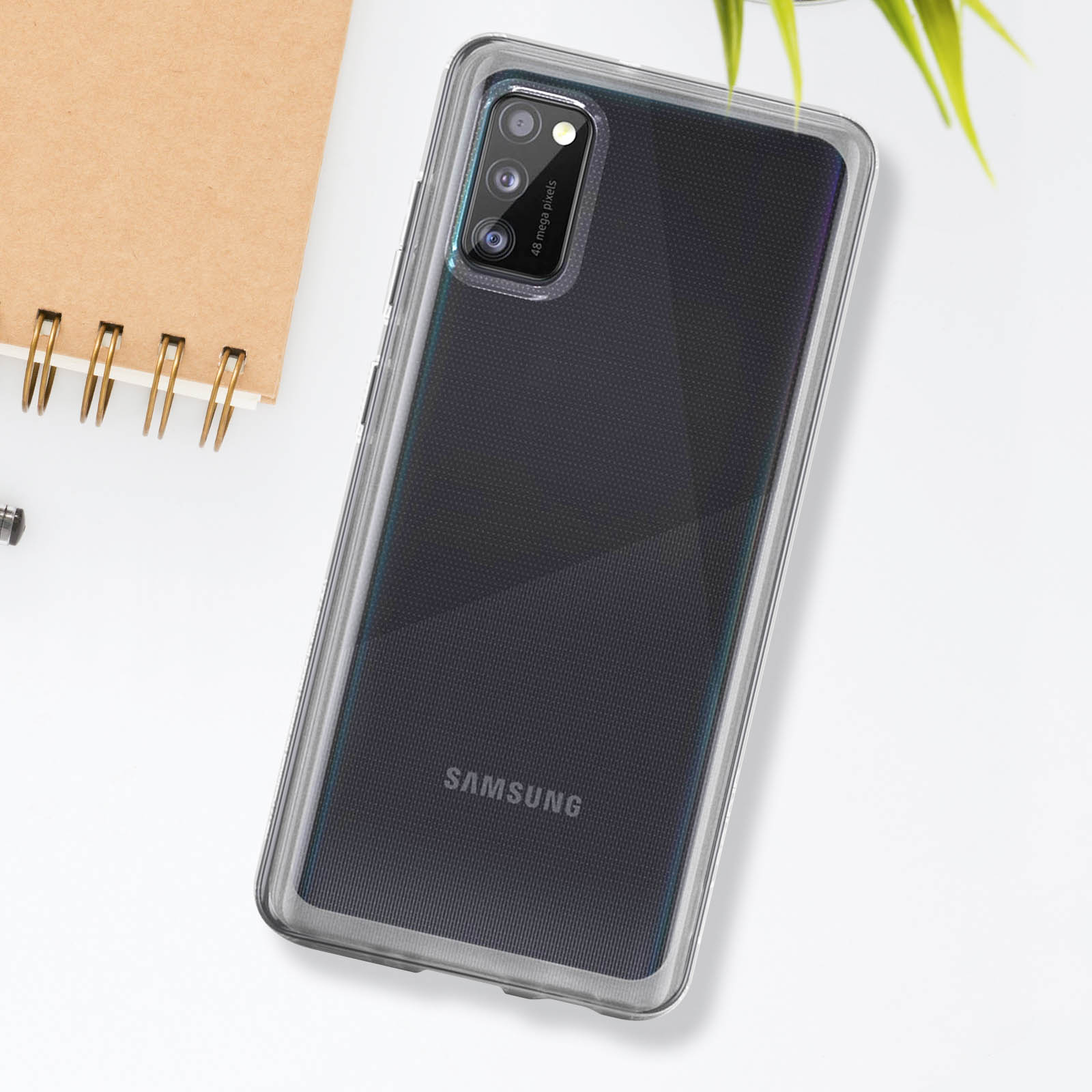 Teléfono en funda transparente sobre superficie blanca. Cuaderno visible. Samsung en el teléfono.