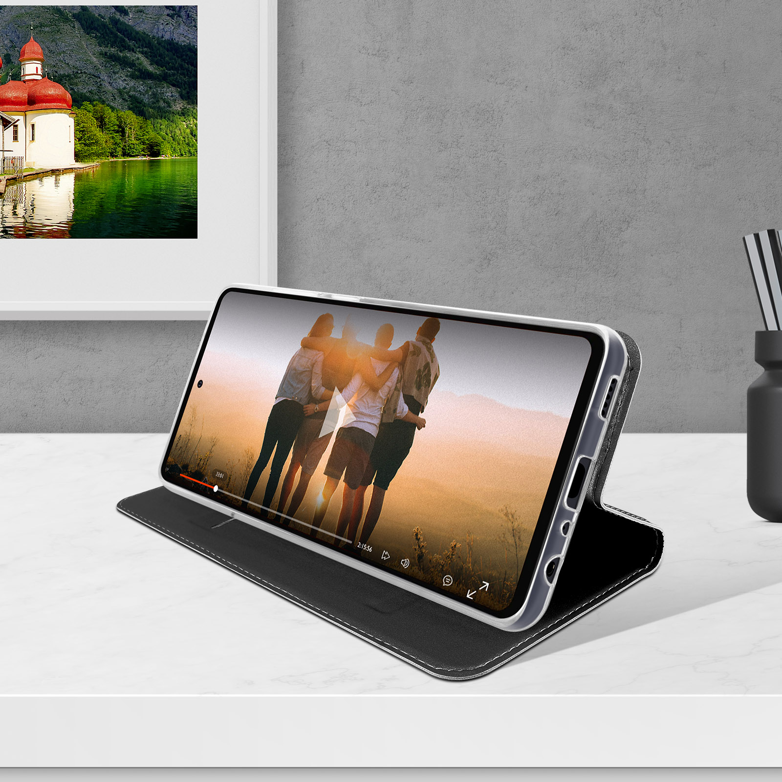 Telefon zeigt Video auf Ständer, weißer Tisch, mit gerahmtem Bild im Hintergrund.