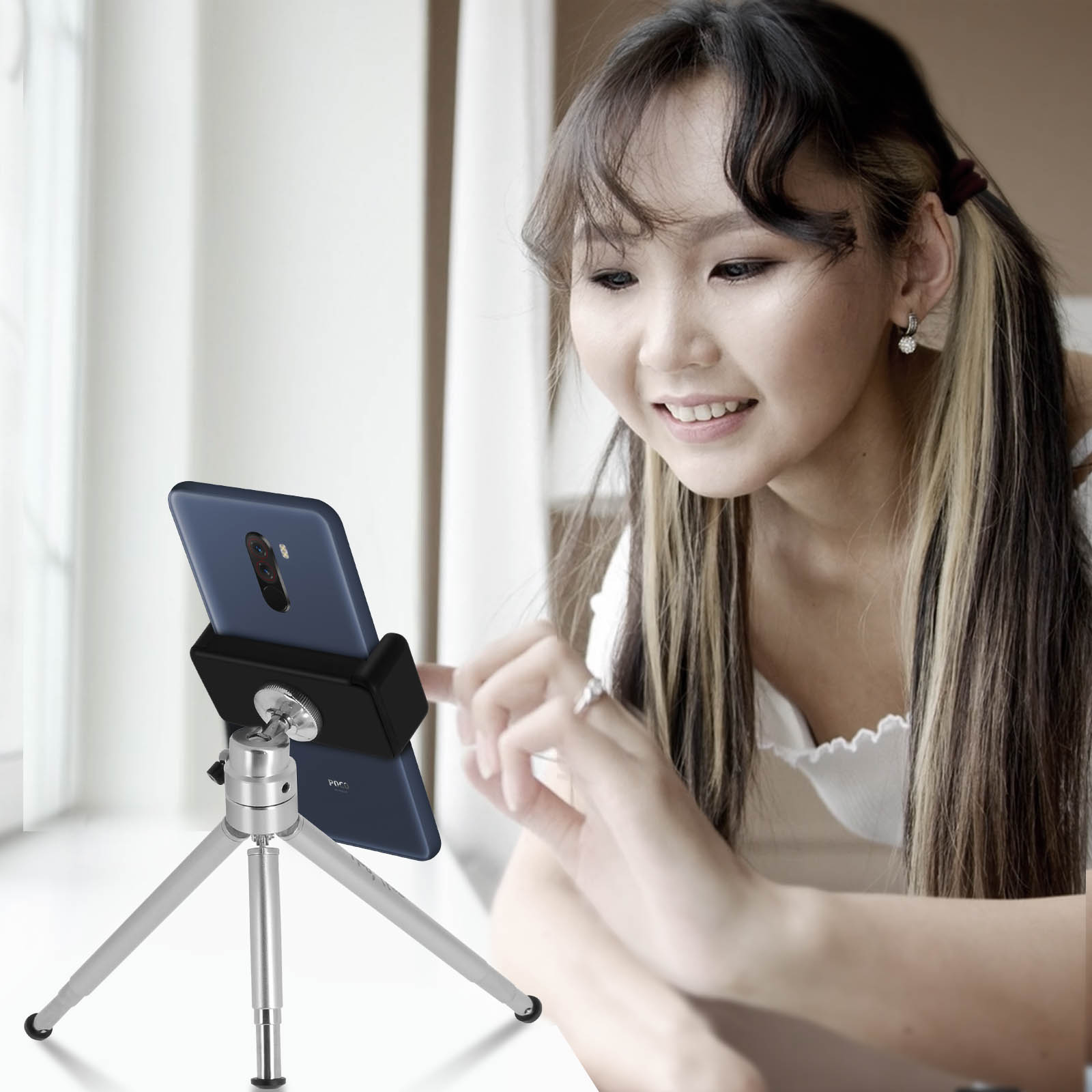 Mujer con teléfono en trípode, sonriendo, luz brillante de ventana. Teléfono mostrando app.