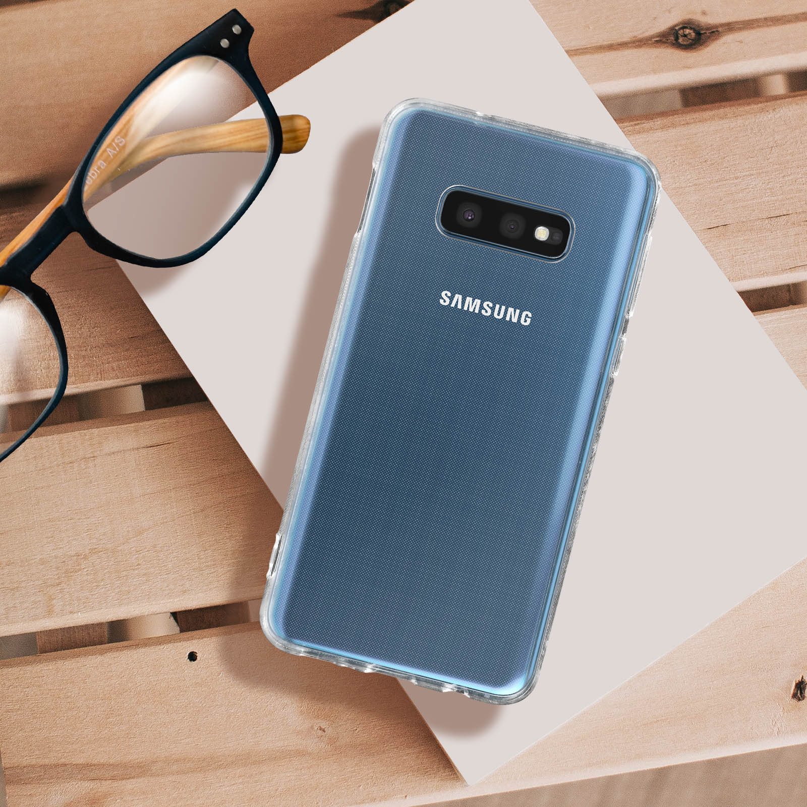 Un telefono in una custodia trasparente è appoggiato su una superficie di legno con occhiali e carta. Logo Samsung.