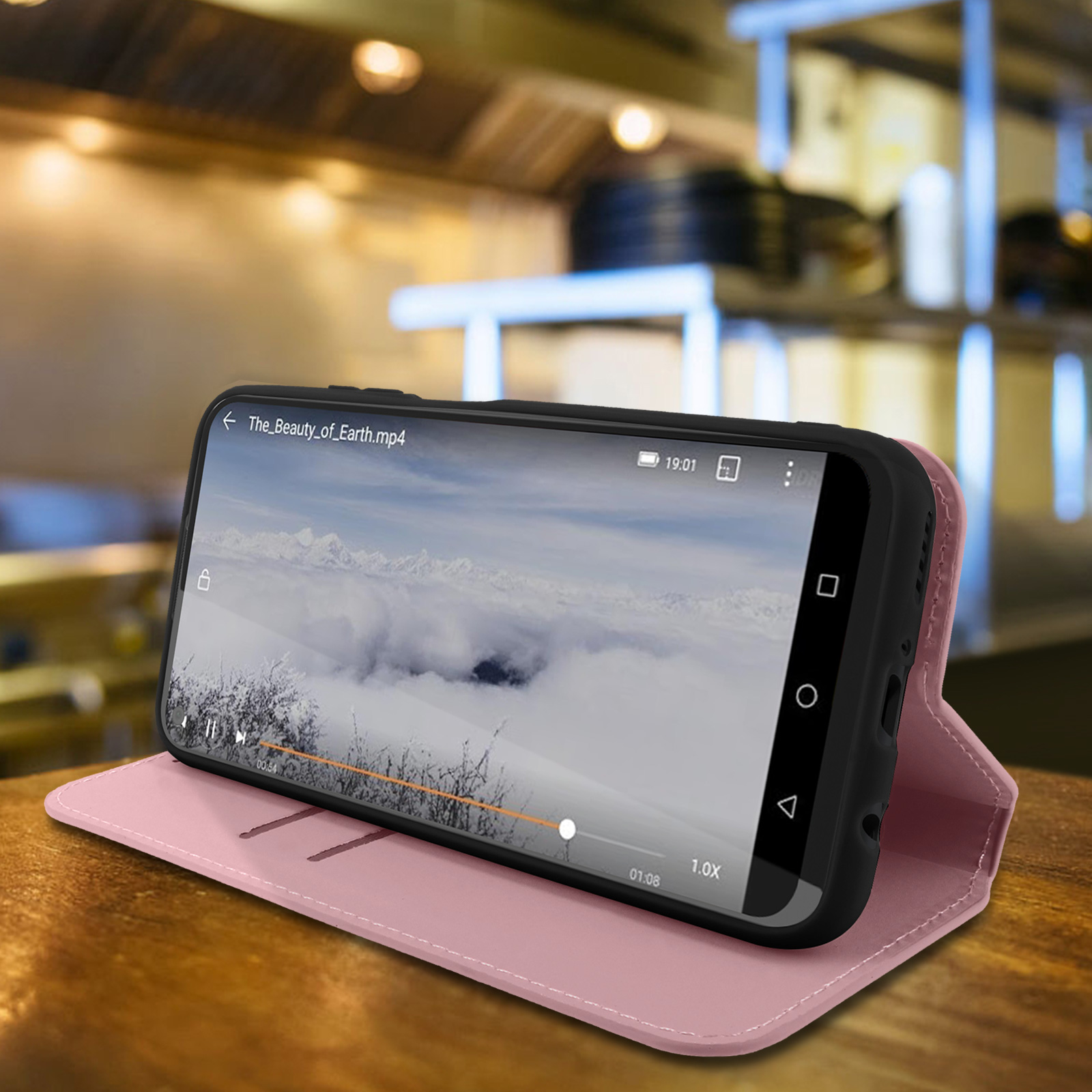 Een telefoon in een roze hoes speelt een video af. De scène is een houten tafel met een wazige achtergrond van een café.