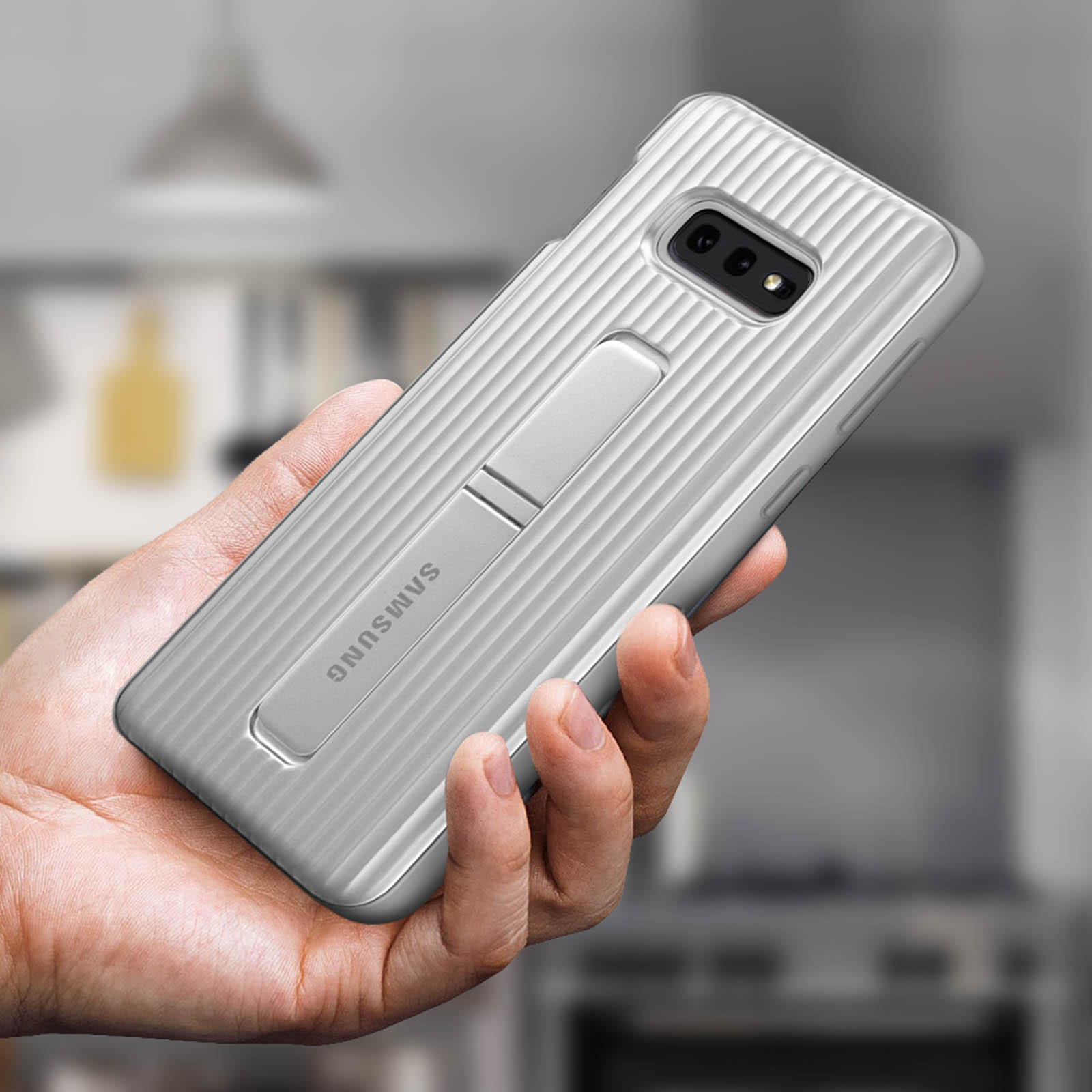 Hand hält silberne Handyhülle mit Ständer, Samsung-Logo sichtbar. Hintergrund ist unscharf.