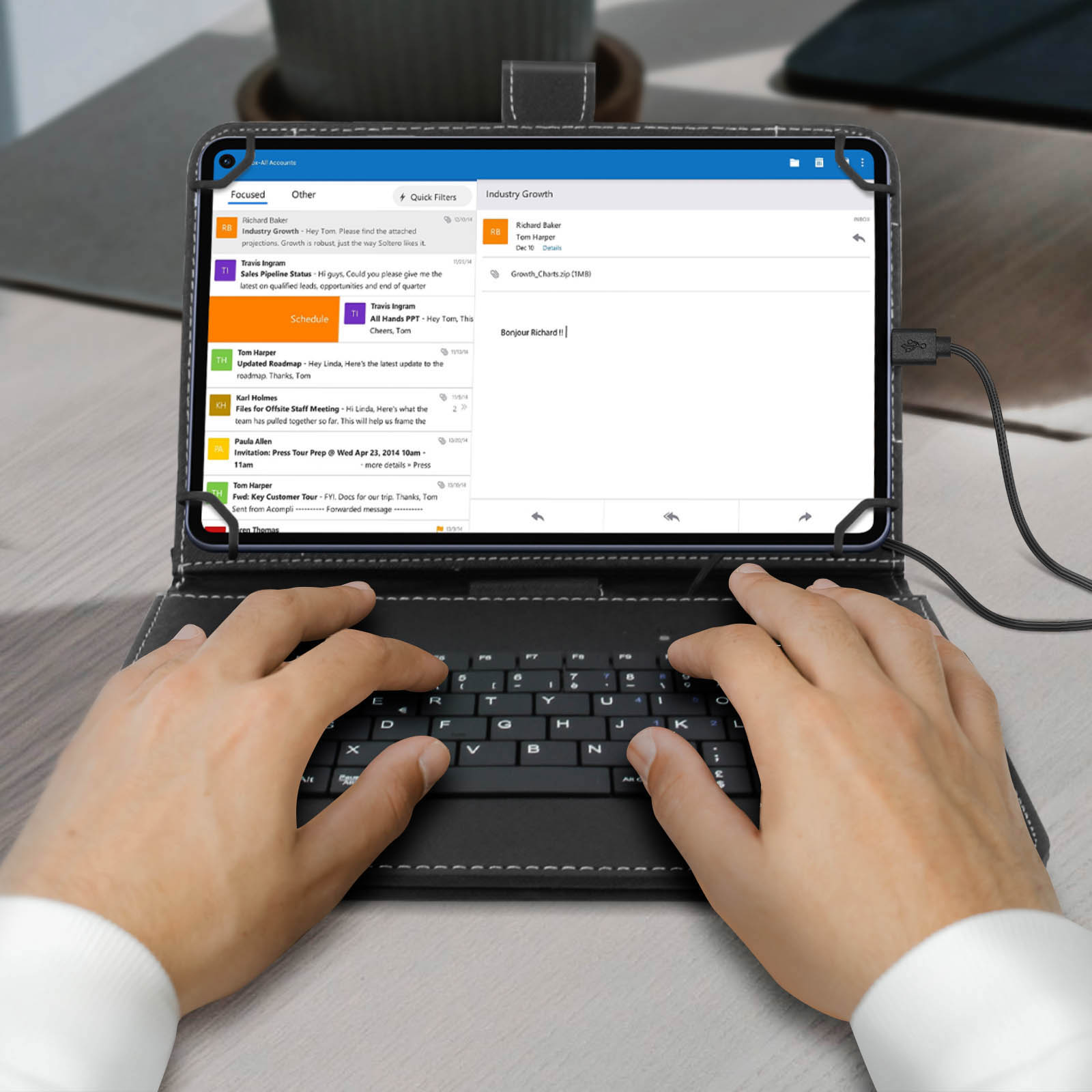 Tableta con funda de teclado, mostrando correos electrónicos. Manos escribiendo en el teclado. Un cable de carga conectado.