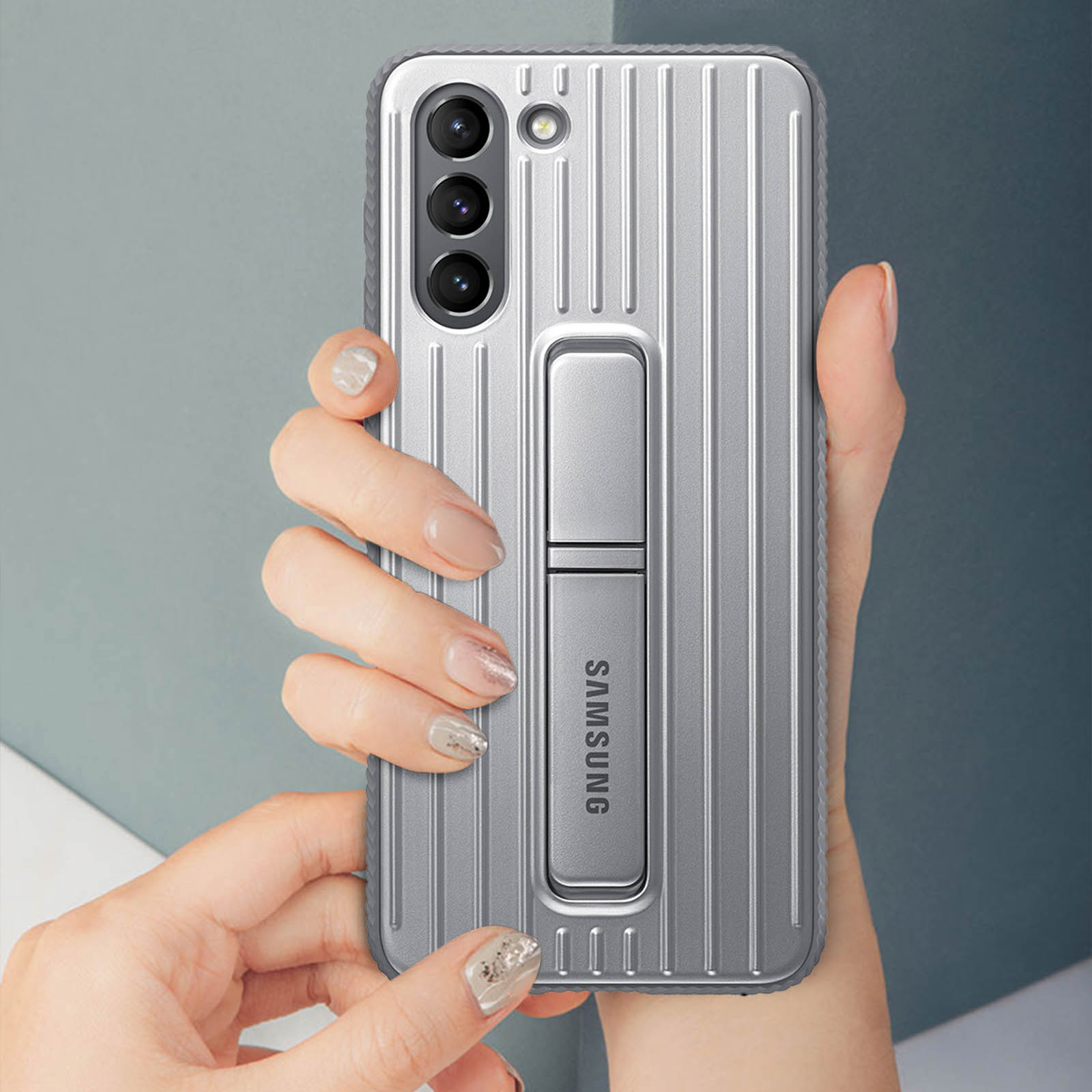 Die Hand einer Person, die eine silberne Samsung-Handyhülle mit vertikalen Linien und einem Ständer hält.