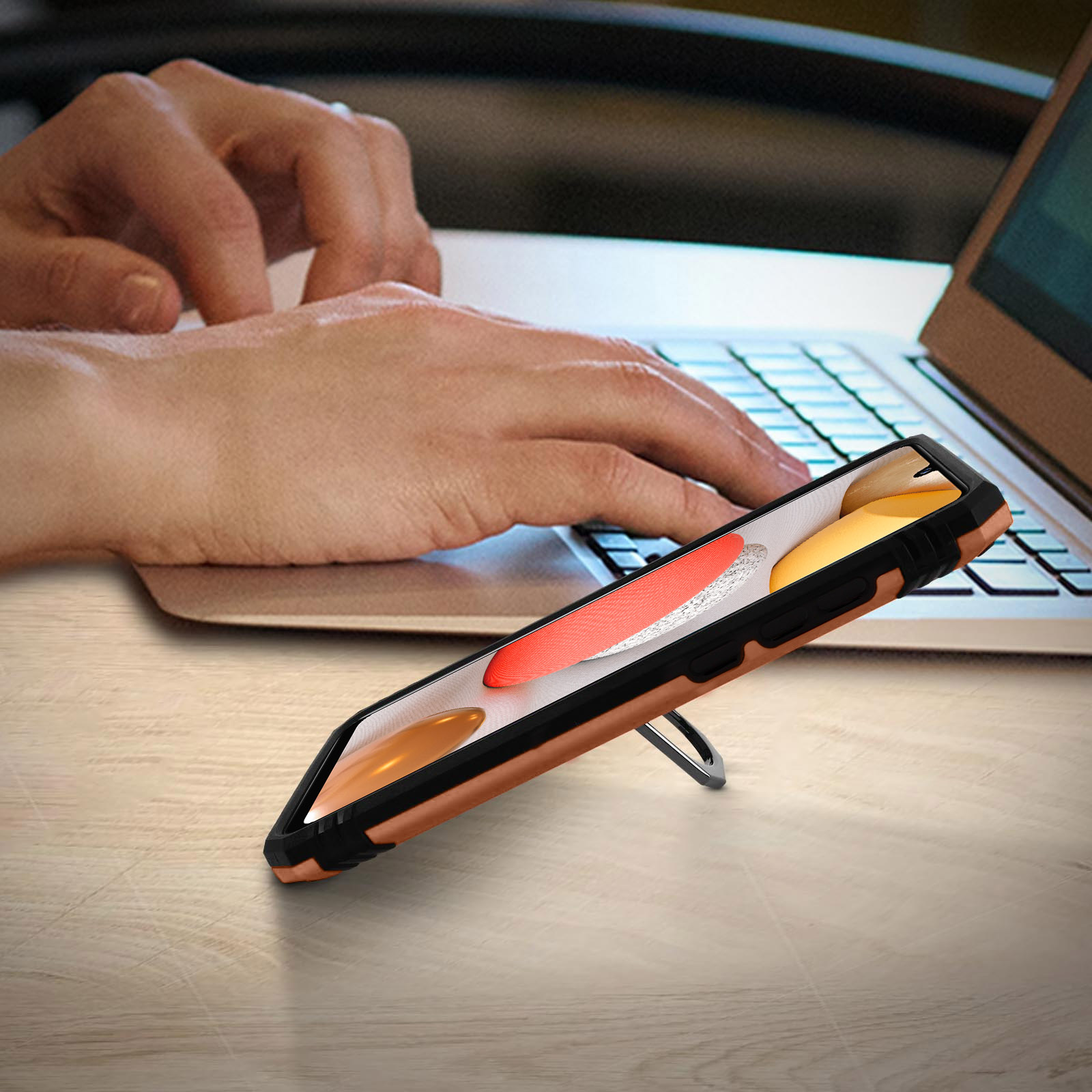 Telefon in Hülle auf Holztisch, Hände tippen im Hintergrund auf Laptop.