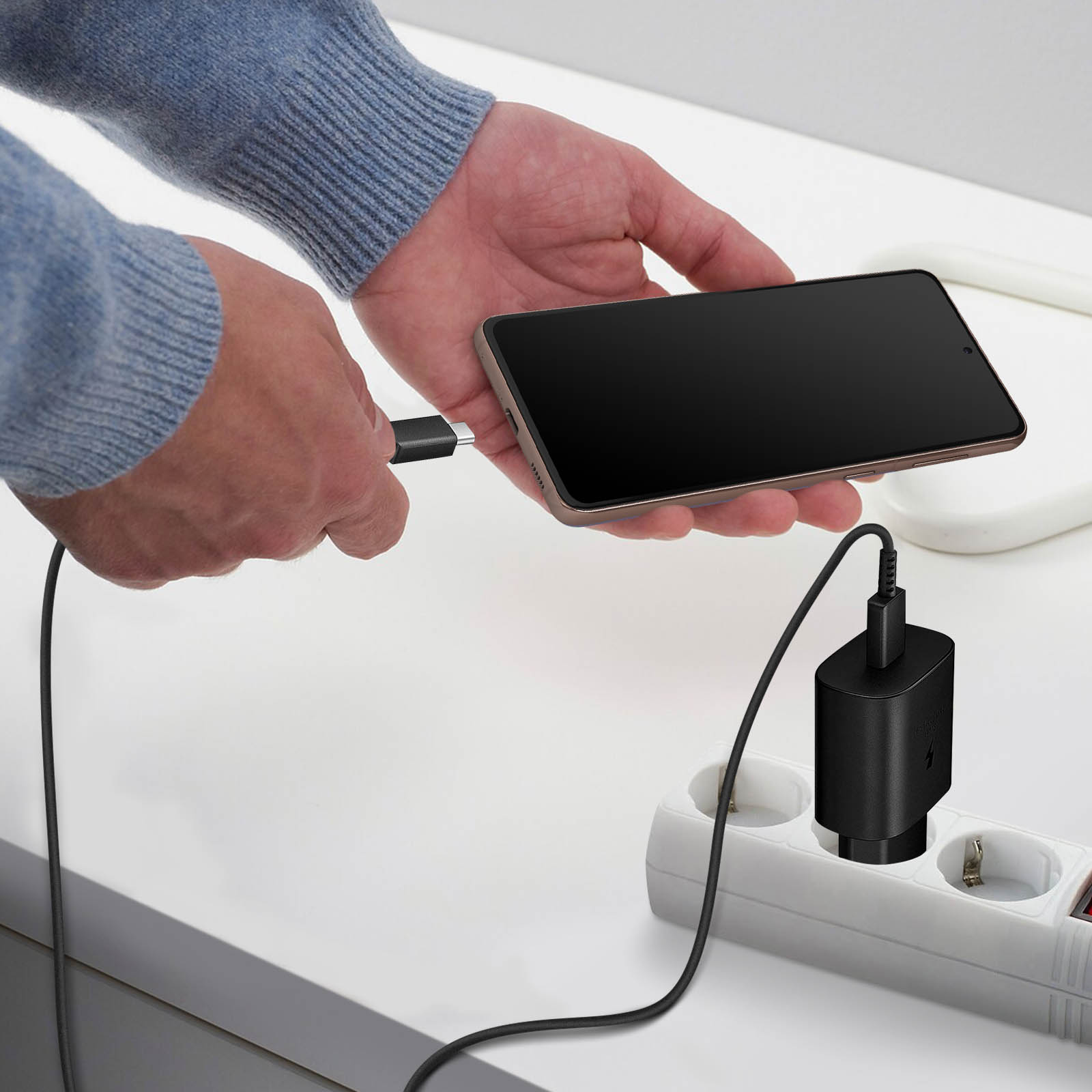 Persona che carica un telefono. Il telefono è collegato a un caricatore collegato a una presa multipla su un tavolo bianco.