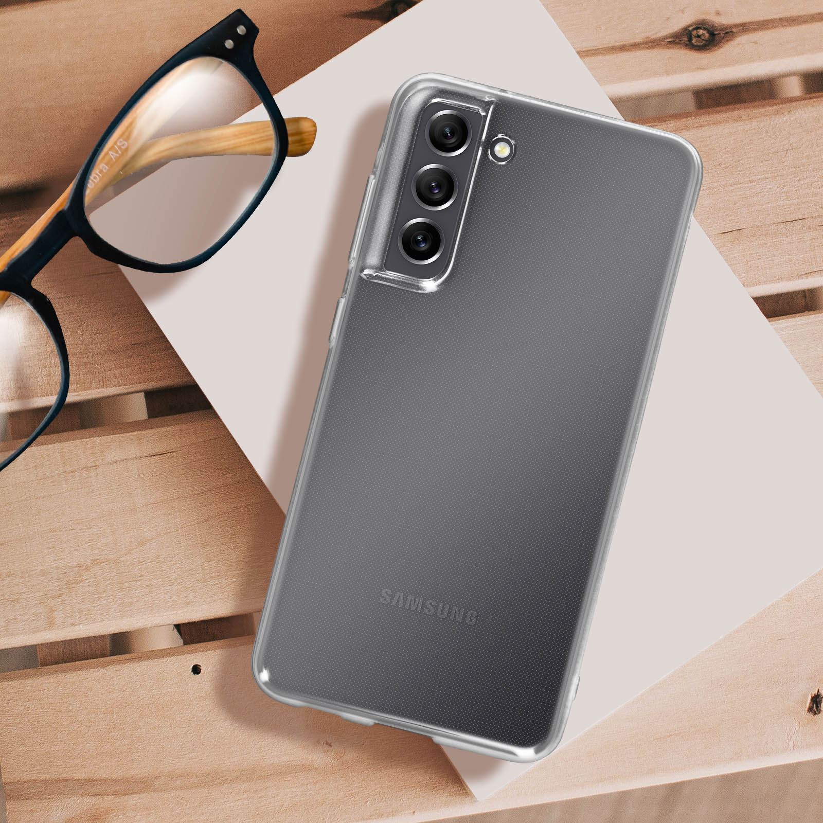 Transparente Handyhülle auf Holztisch mit Brille. Samsung-Handy und weißes Papier daneben.