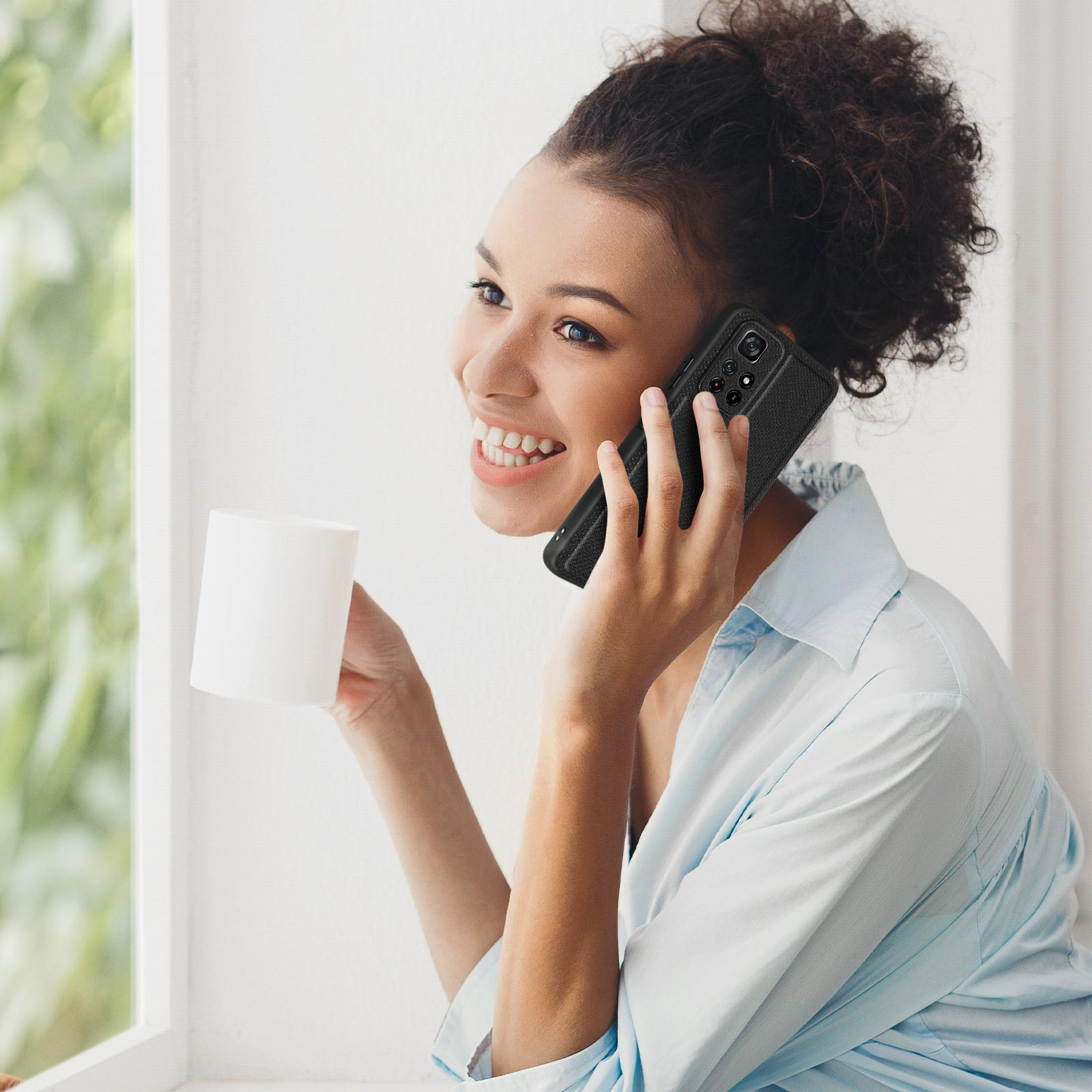 Donna sorridente al telefono, tiene una tazza bianca. È vicino a una finestra.
