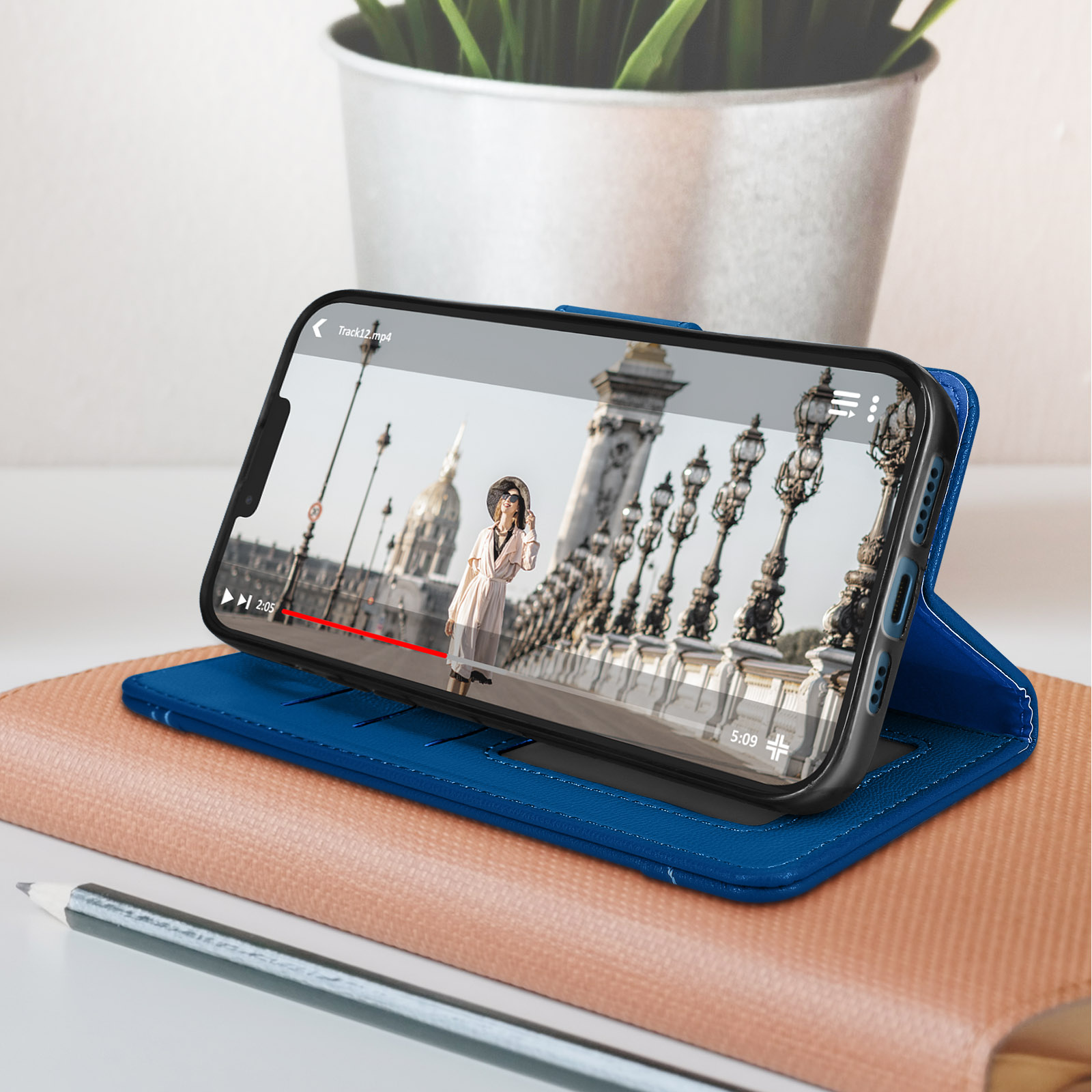 Téléphone dans un étui bleu, diffusant une vidéo d'un pont. Cahier beige et crayon au premier plan.