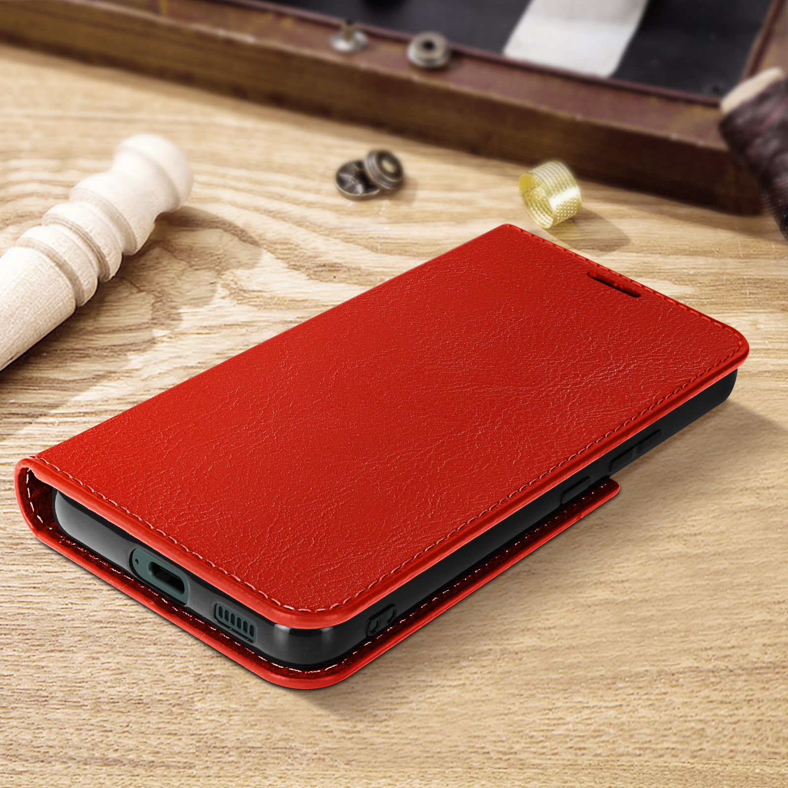 Custodia per telefono in pelle rossa aperta su un tavolo di legno. Sullo sfondo, accessori da cucito.