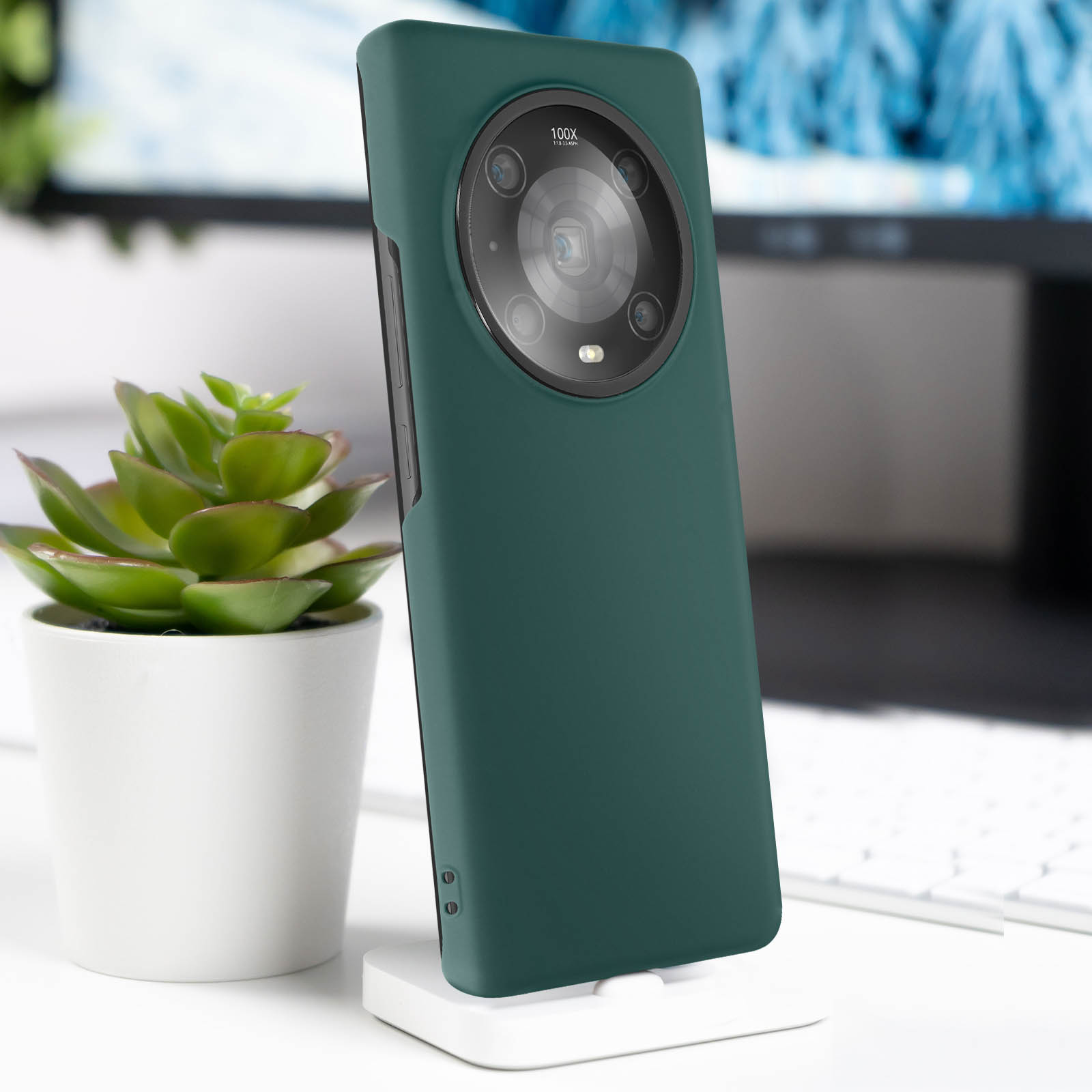 Custodia verde su un supporto. La custodia ha un ampio modulo fotocamera. Scrivania bianca e pianta.