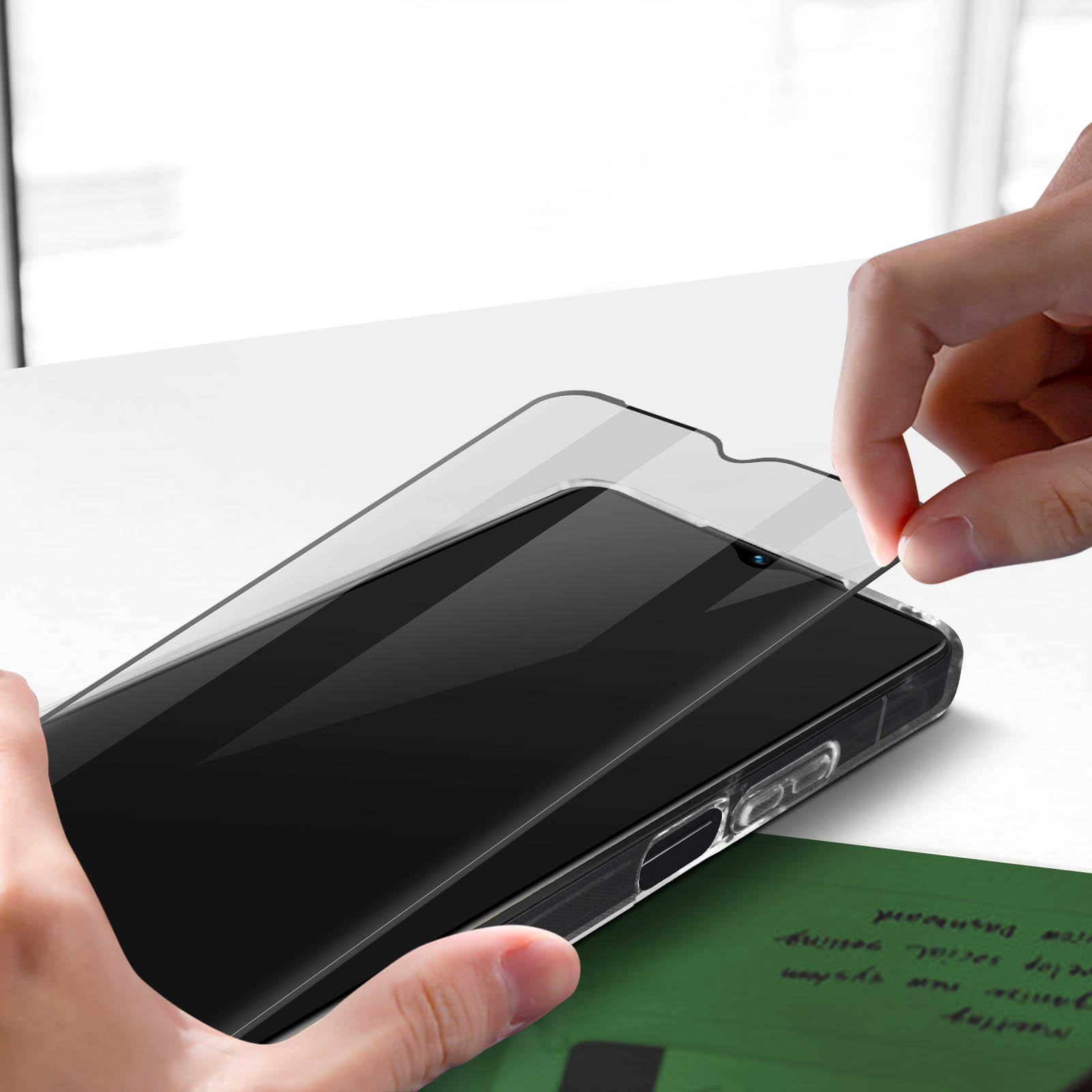 Hand legt Displayschutz auf Telefon. Klares Glas über schwarzem Telefon. Weißer Tisch und grünes Papier rechts.