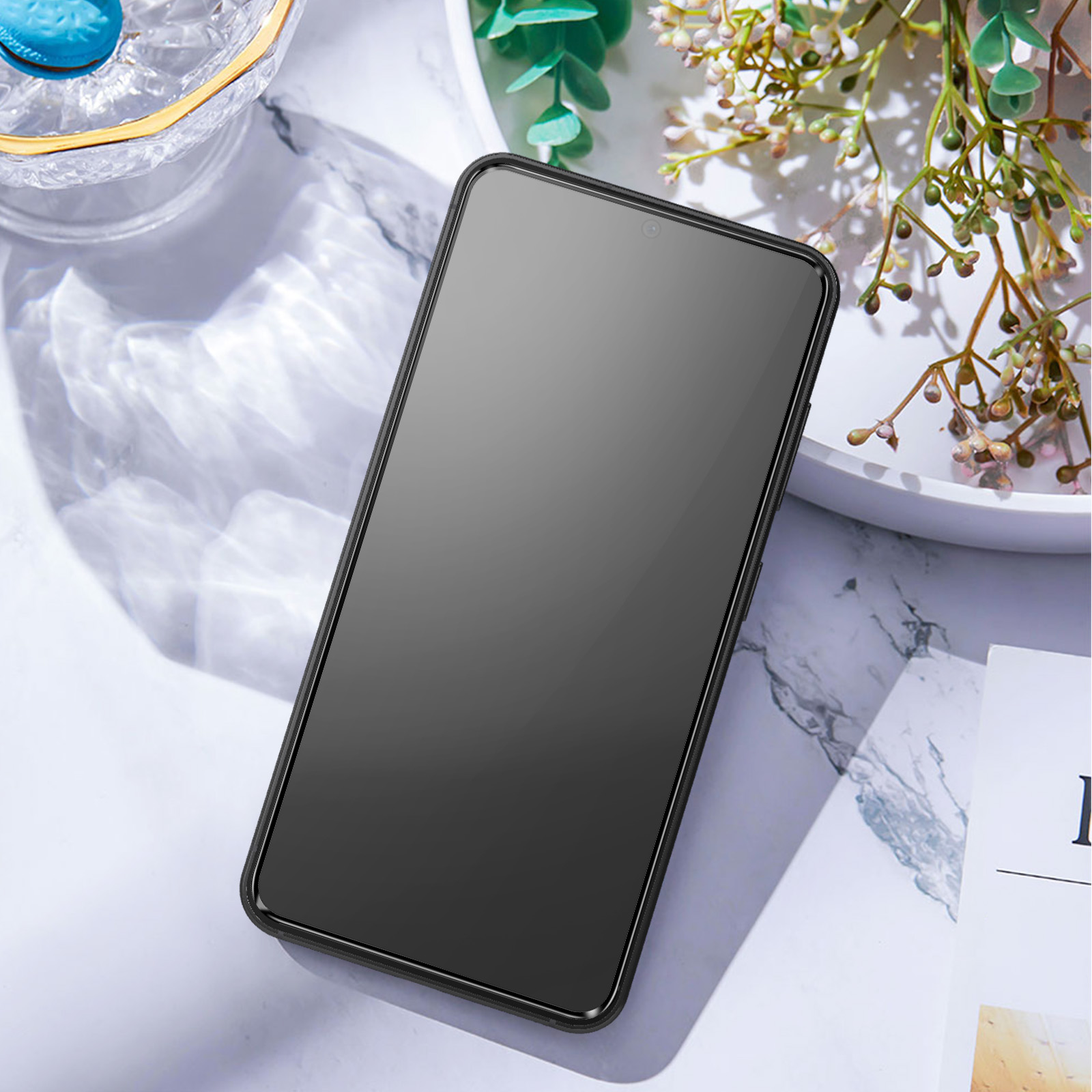 Un téléphone noir avec un écran sombre est sur une surface blanche, à côté d'un vase avec des plantes.