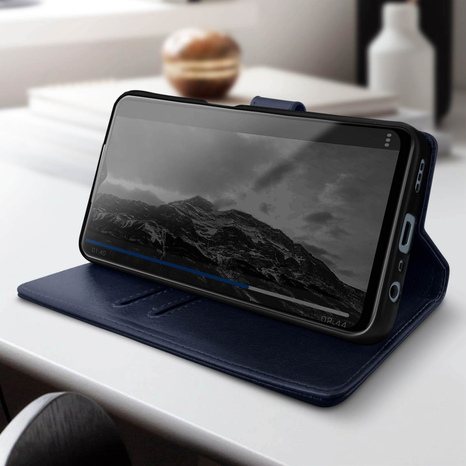 Telefono in custodia blu scuro, mostra un video di un paesaggio montano. Su una superficie bianca, con oggetti sullo sfondo.