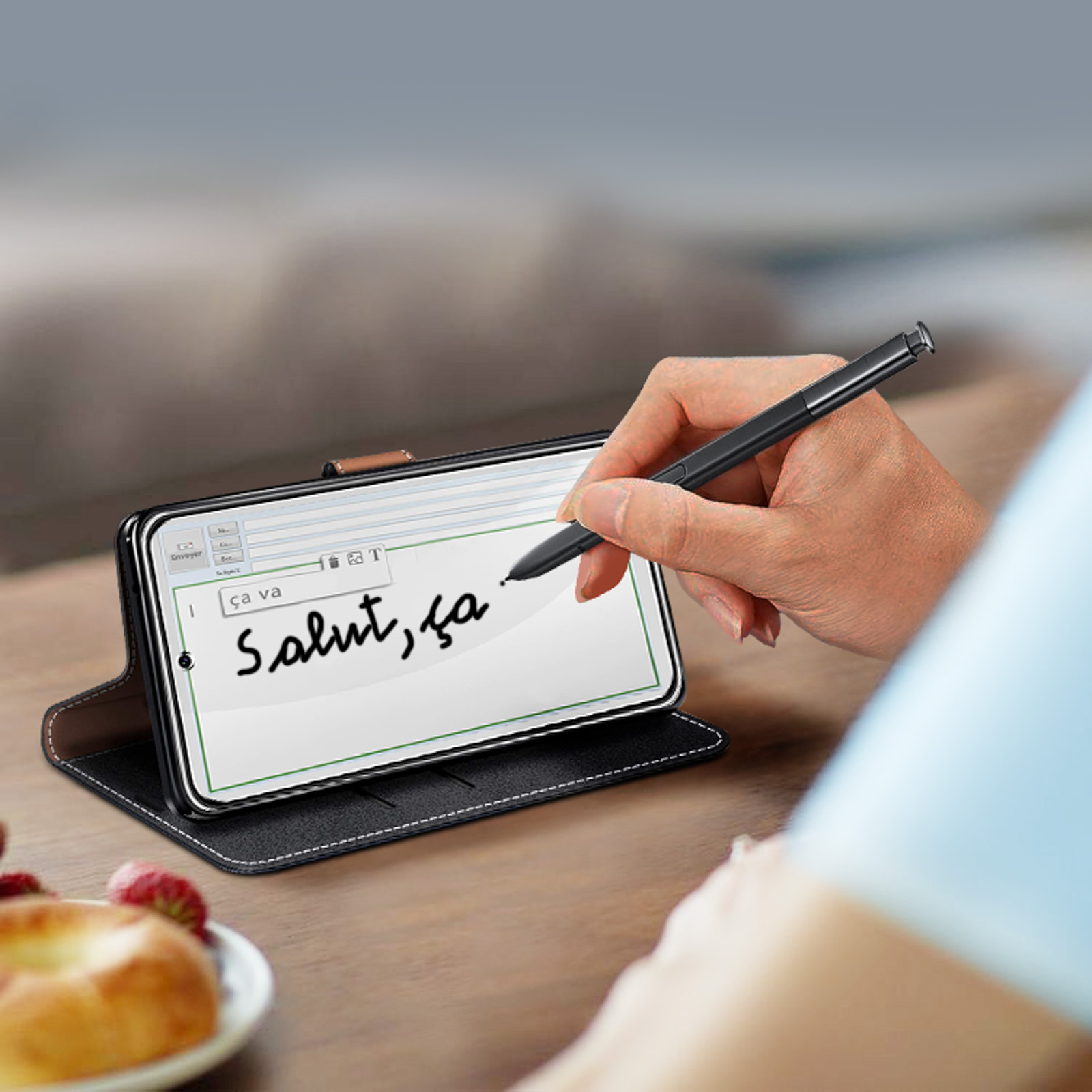 Una mano escribiendo en la pantalla de un teléfono con un lápiz óptico. La pantalla muestra un mensaje. Un bagel está en el fondo.