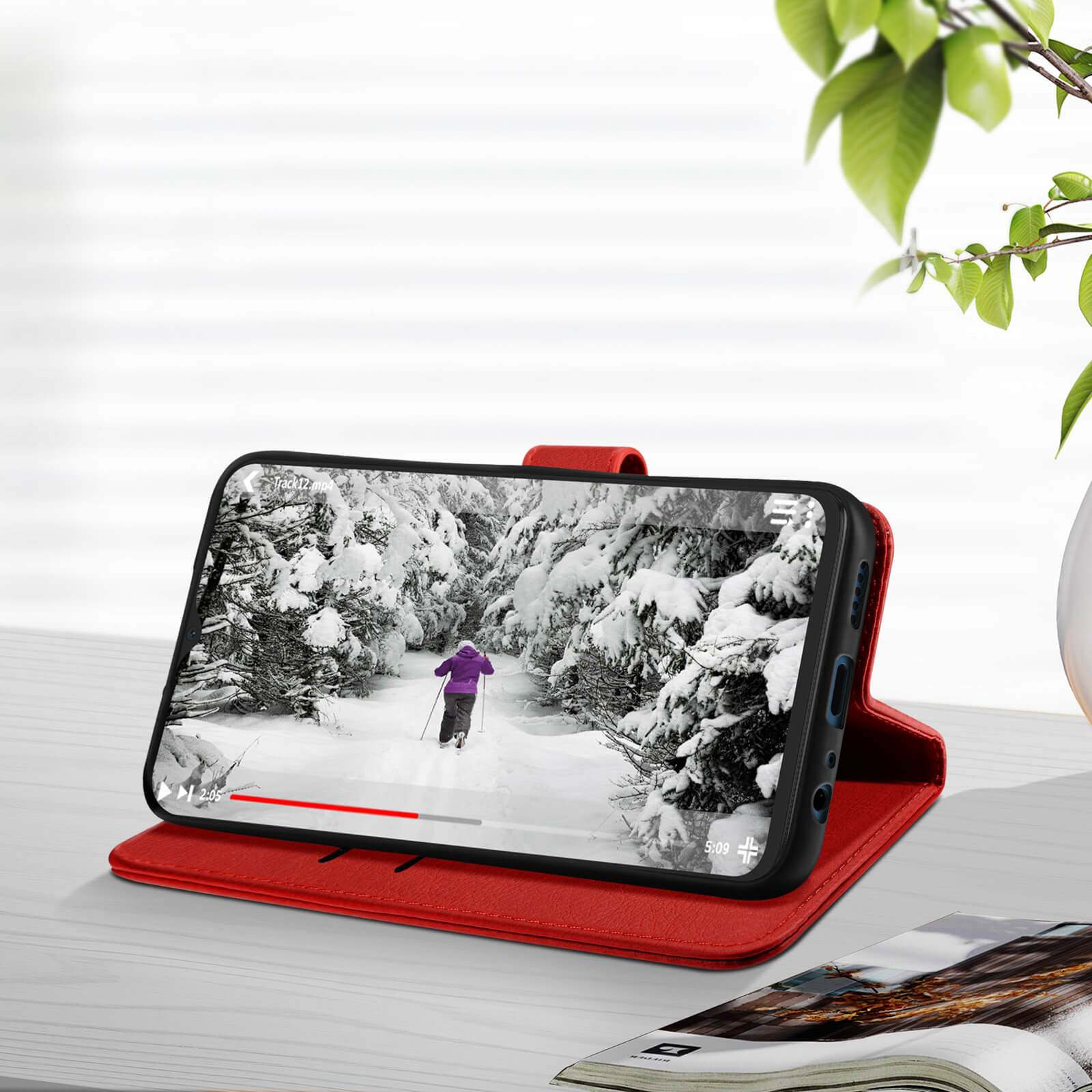 Ein Telefon zeigt eine verschneite Szene. Es ist in einer roten Hülle auf einem Tisch mit einer Pflanze im Hintergrund.