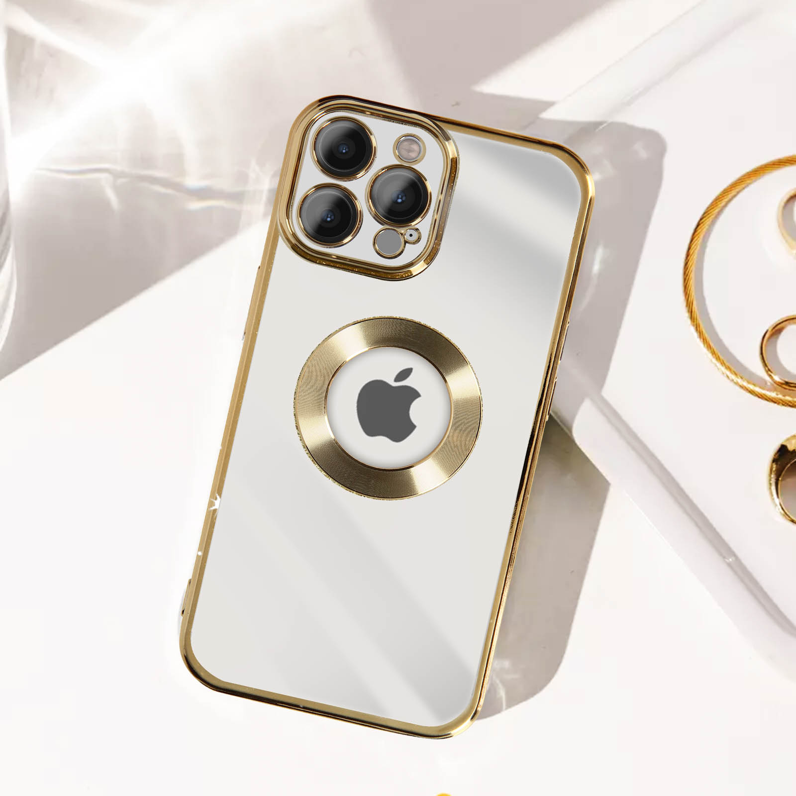 Custodia bianca per telefono con finiture dorate e logo Apple, su una superficie bianca con gioielli.