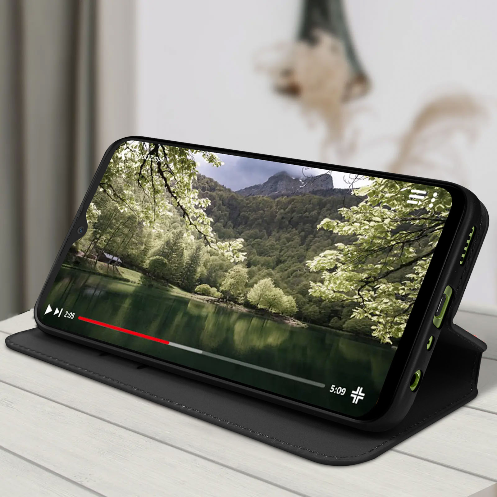 Smartphone in een zwarte portemonneehoes, die een video van een meer en bomen afspeelt. Een houten tafel staat op de achtergrond.
