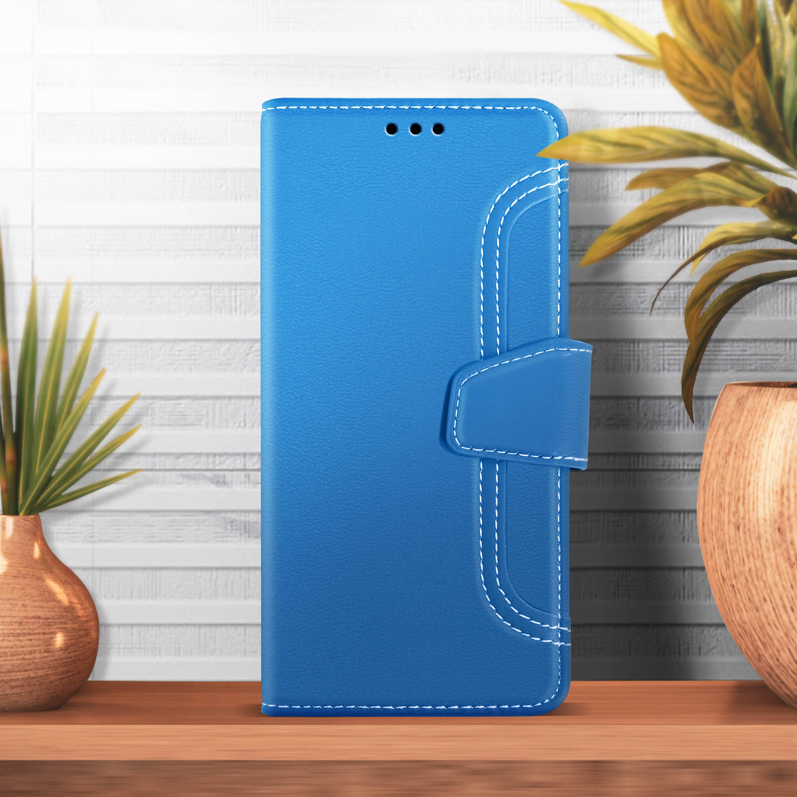 Custodia per telefono in pelle blu. Cuciture bianche. Chiusa con un fermaglio. Tavolo in legno, pianta verde in vaso.