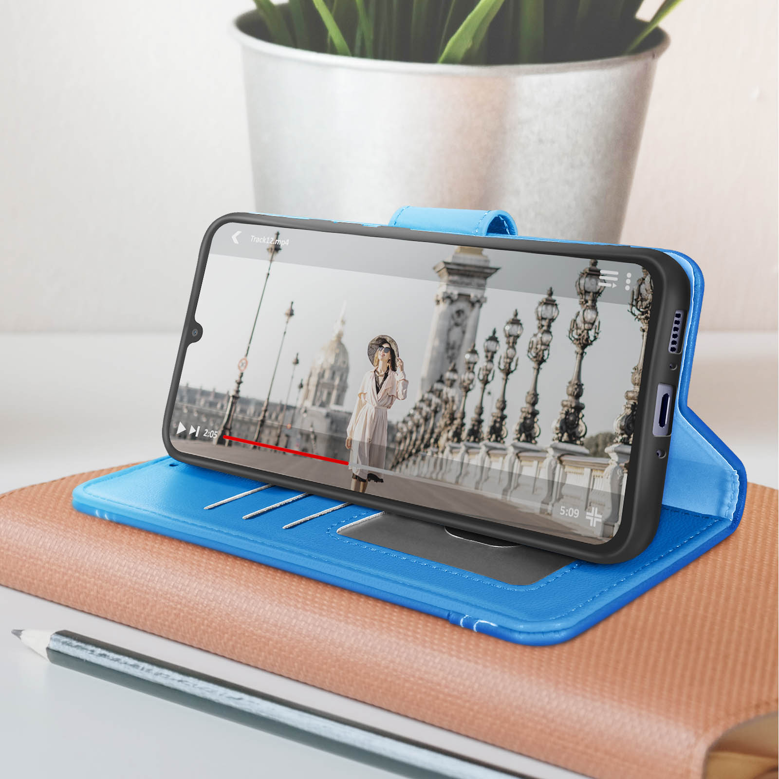 Smartphone in custodia blu che mostra un video. Lo sfondo include una pianta e accessori.