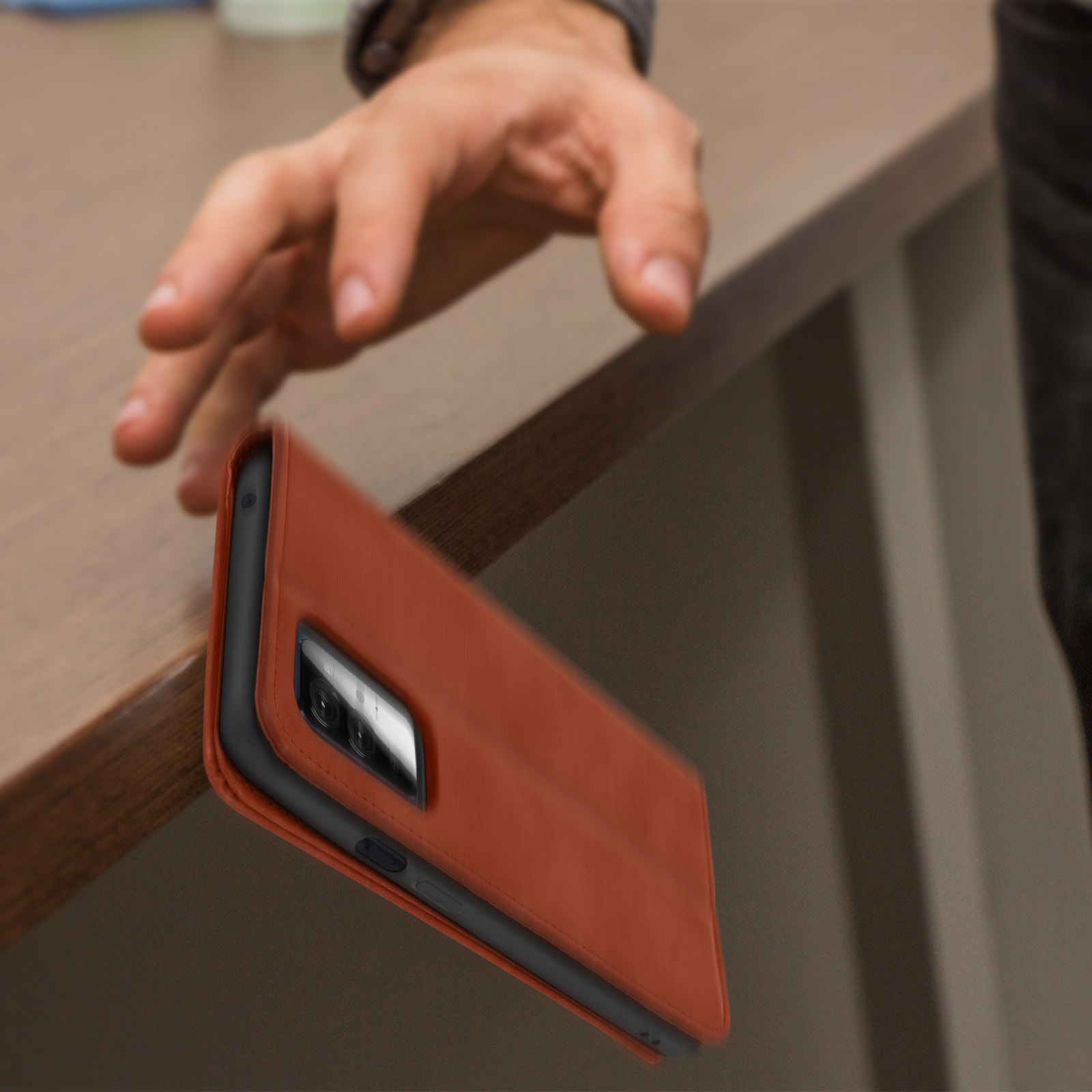Ein Telefon in einer orangefarbenen Hülle fällt von einem Tisch. Eine Hand ist über dem Telefon.
