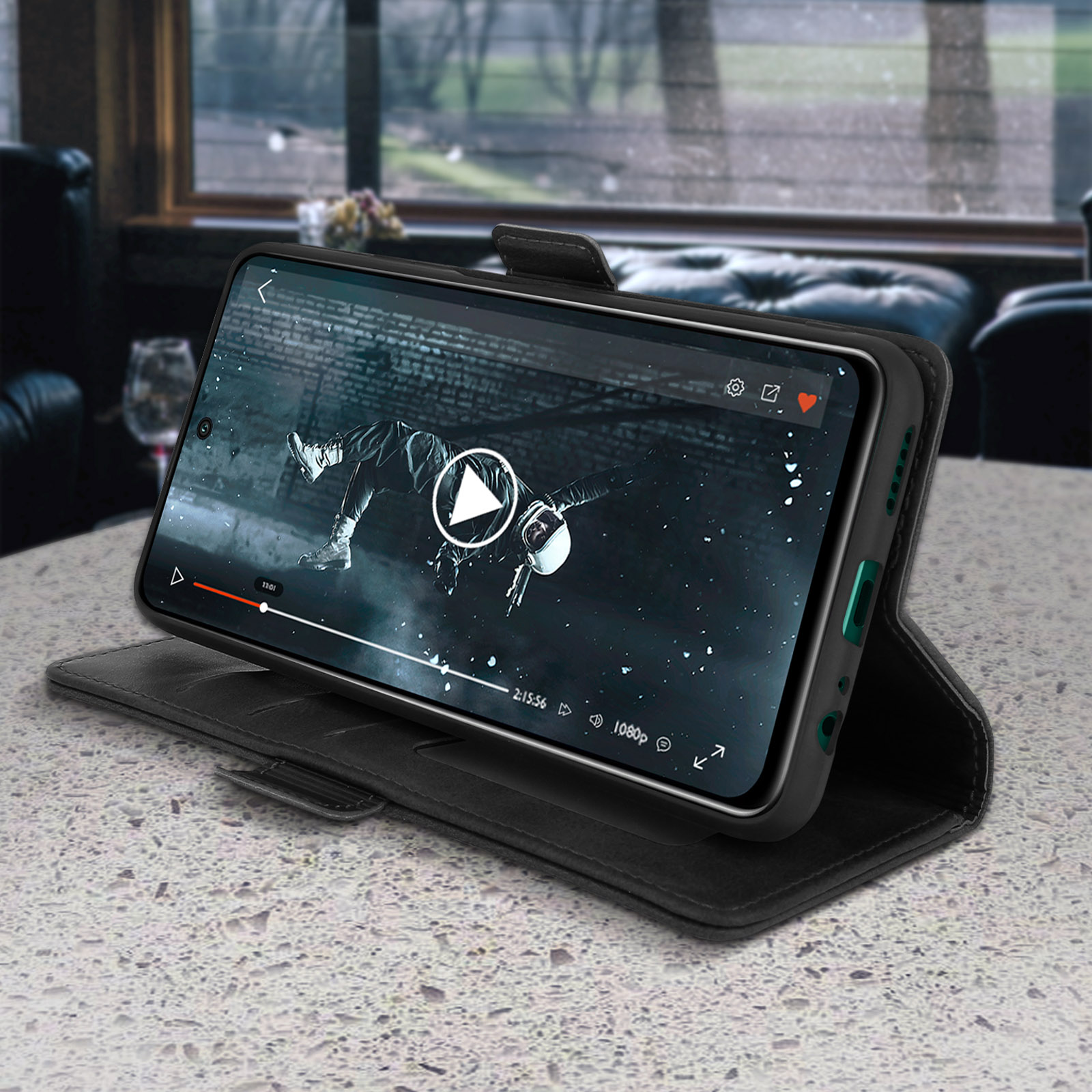 Schwarzes Telefon in einer Hülle auf einem Tisch. Das Telefon zeigt ein Video. Der Hintergrund ist ein dunkler Raum.