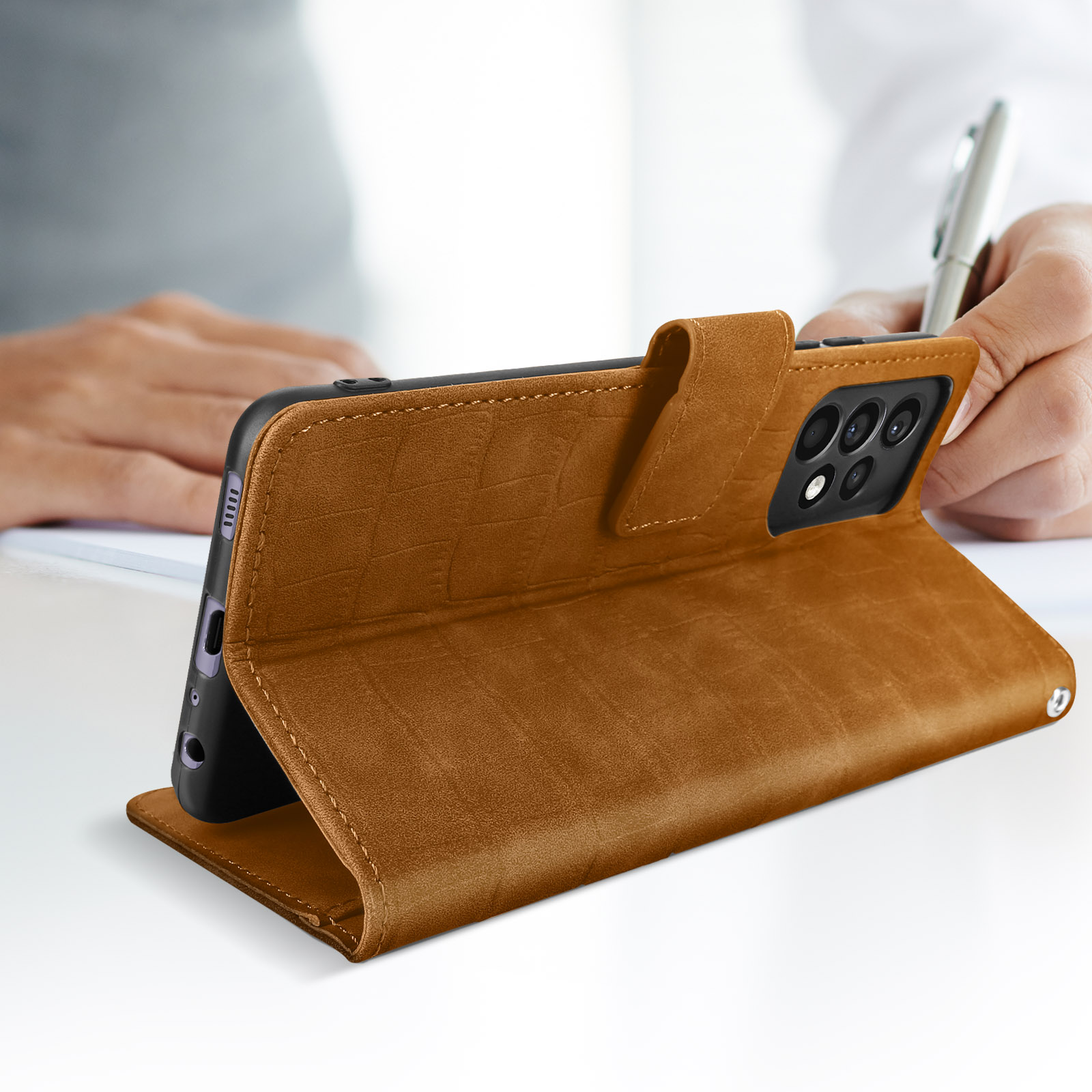 Braune Handyhülle, auf einem Tisch geöffnet. Eine Person schreibt. Telefon ist angewinkelt, offen abgestützt.
