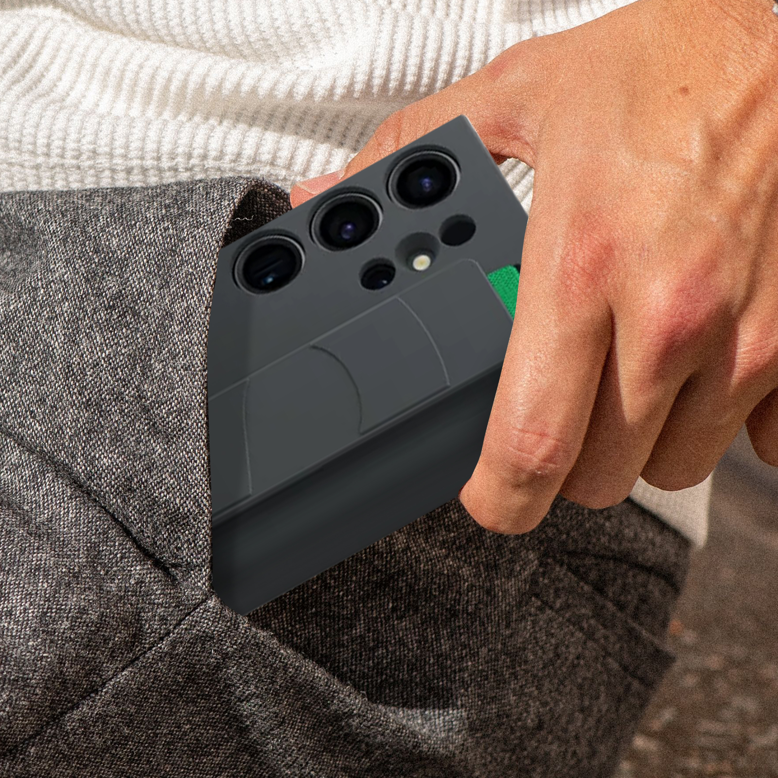 Hand in de zak met een zwarte telefoonhoes. Grijze broek en een vage achtergrond.