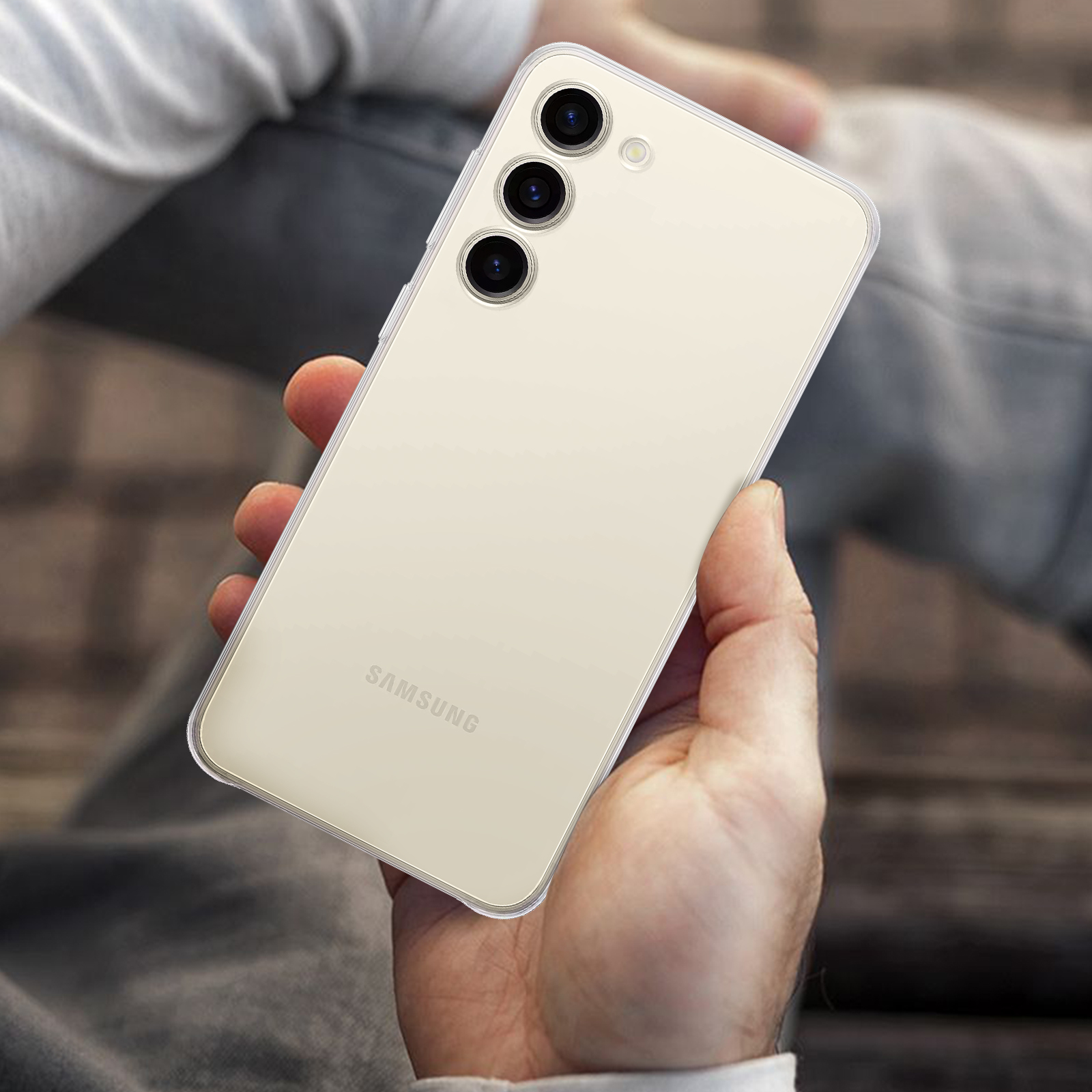 Dłoń trzyma jasny telefon z trzema aparatami. Tłem są kolana osoby.