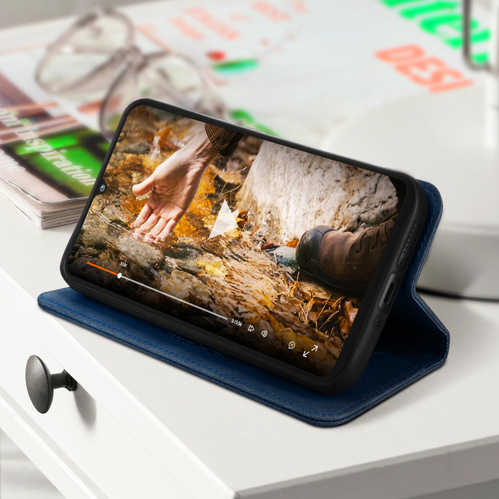 Telefoon in een blauwe hoes, met een video op tafel. Een tijdschrift en een bril liggen in de buurt.